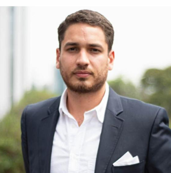 A man in a navy blazer and white shirt standing outdoors with greenery in the background.