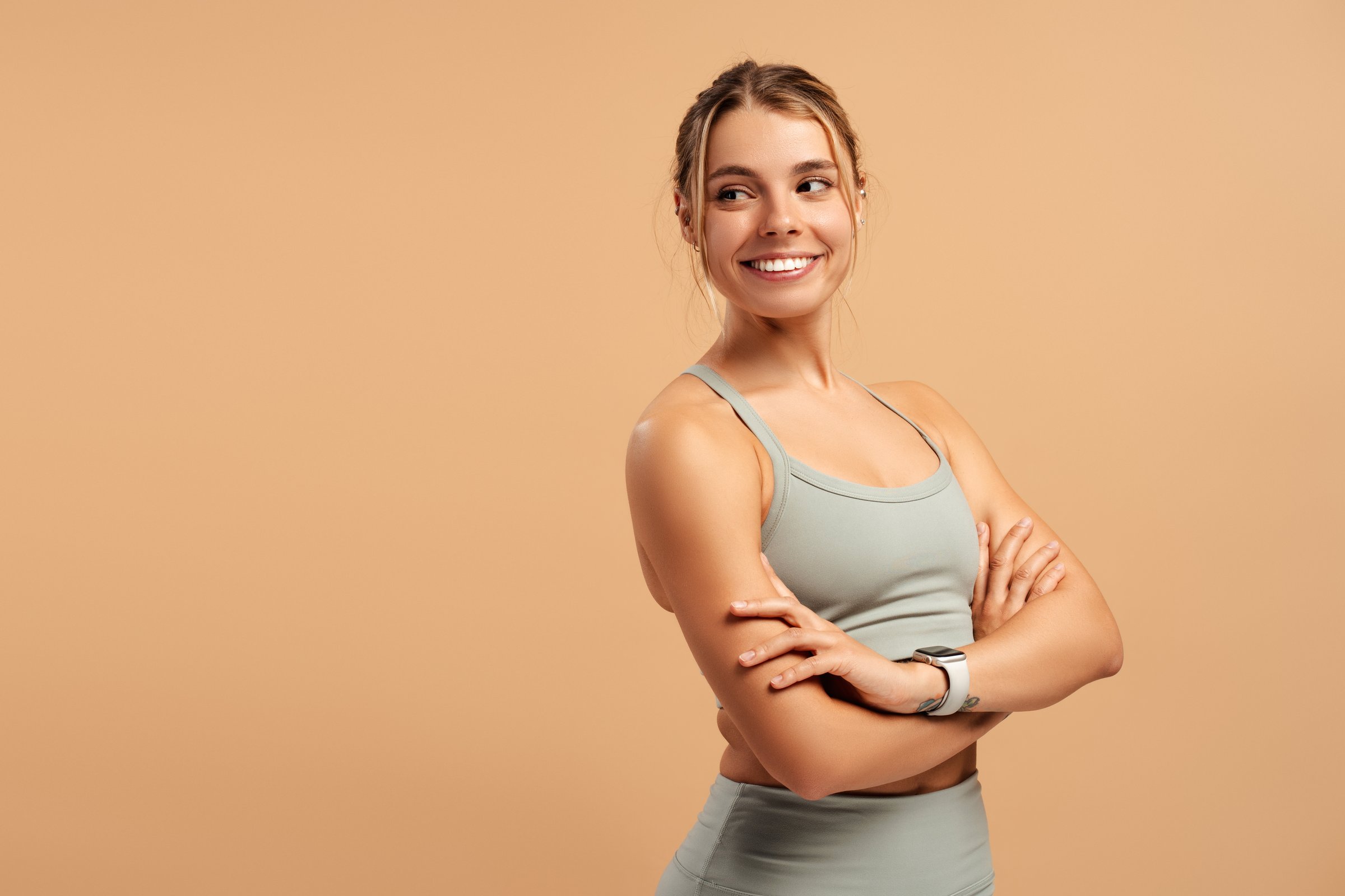 Fit smiling woman in sportswear with arms crossed looking at copy space in studio, isolated on beige background. Sport, health care concept