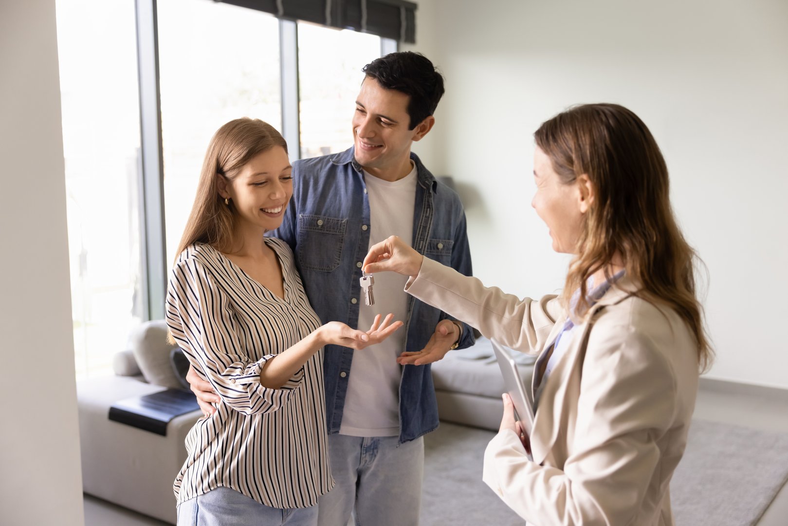 Real estate agent woman giving key to young couple of hugging renters. Happy husband and wife receiving house. New property owners meeting with real estate agent in accommodation for rent