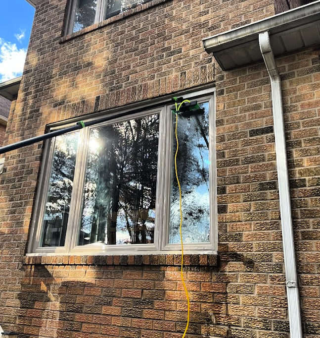 Cleaning brush on a pole washing a large window on a brick house reflecting trees and sky.