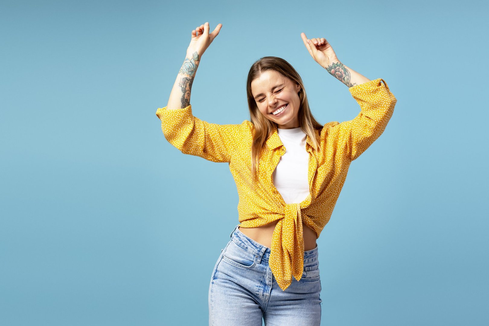 Cheerful young woman with tattoos is dancing with her arms raised and eyes closed, wearing a stylish yellow shirt and jeans against a vibrant blue backdrop, expressing joy and freedom