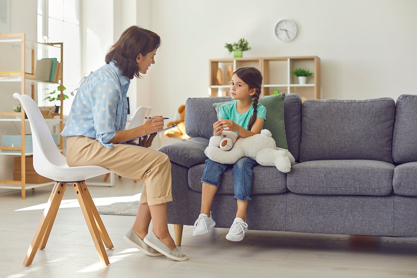Professional female child psychologist working with a little preschool girl in a bright office. Woman makes notes about the condition of the girl on paper. Children's mental therapy