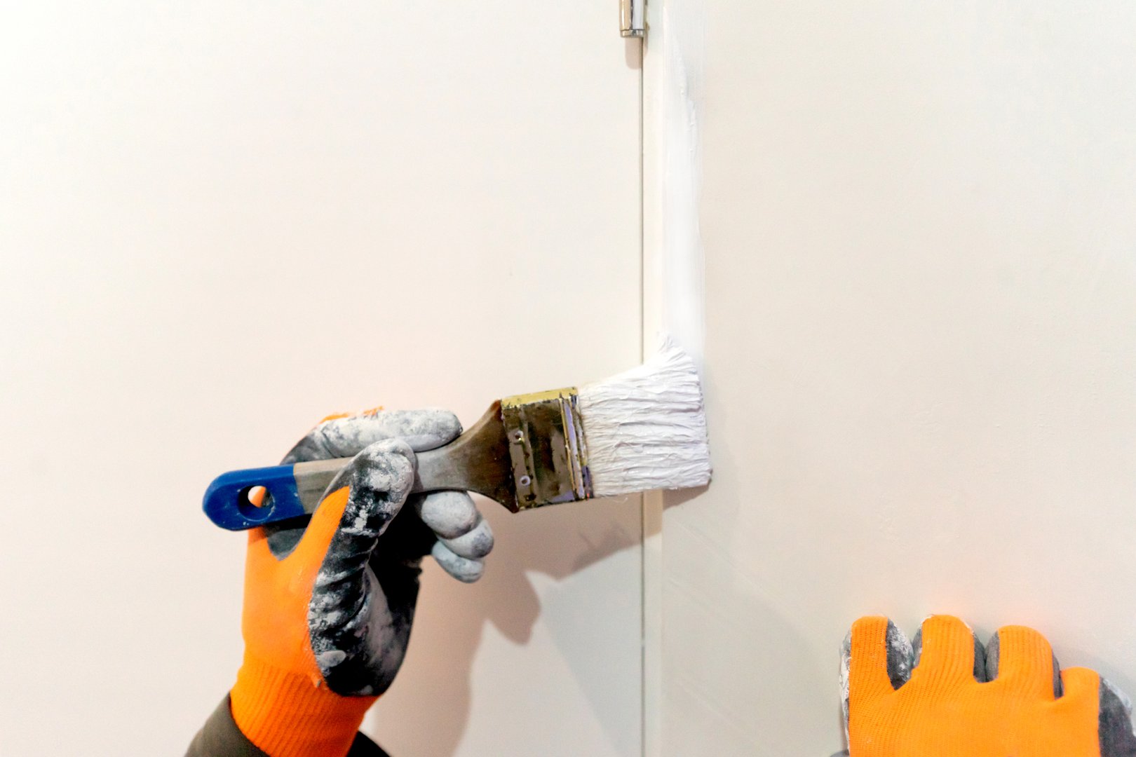 A worker with gloves paints the edge of a door with a brush using white paint. Home repair and improvement. Construction tools and materials, painting work