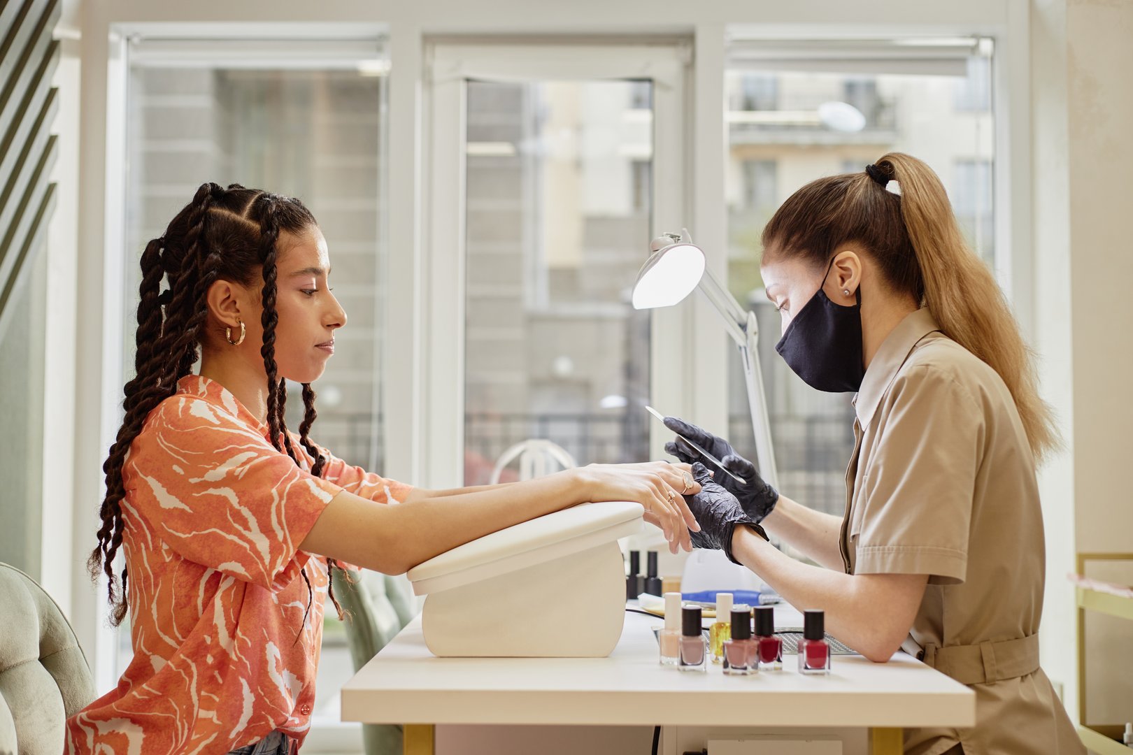 Caucasian young adult woman wearing protective mask performing manicure for biracial young adult woman sitting at nail salon table, both focused on hands, nail polish bottles visible