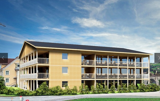 Modern three-story building with wooden exterior and balconies, surrounded by greenery under a clear blue sky.