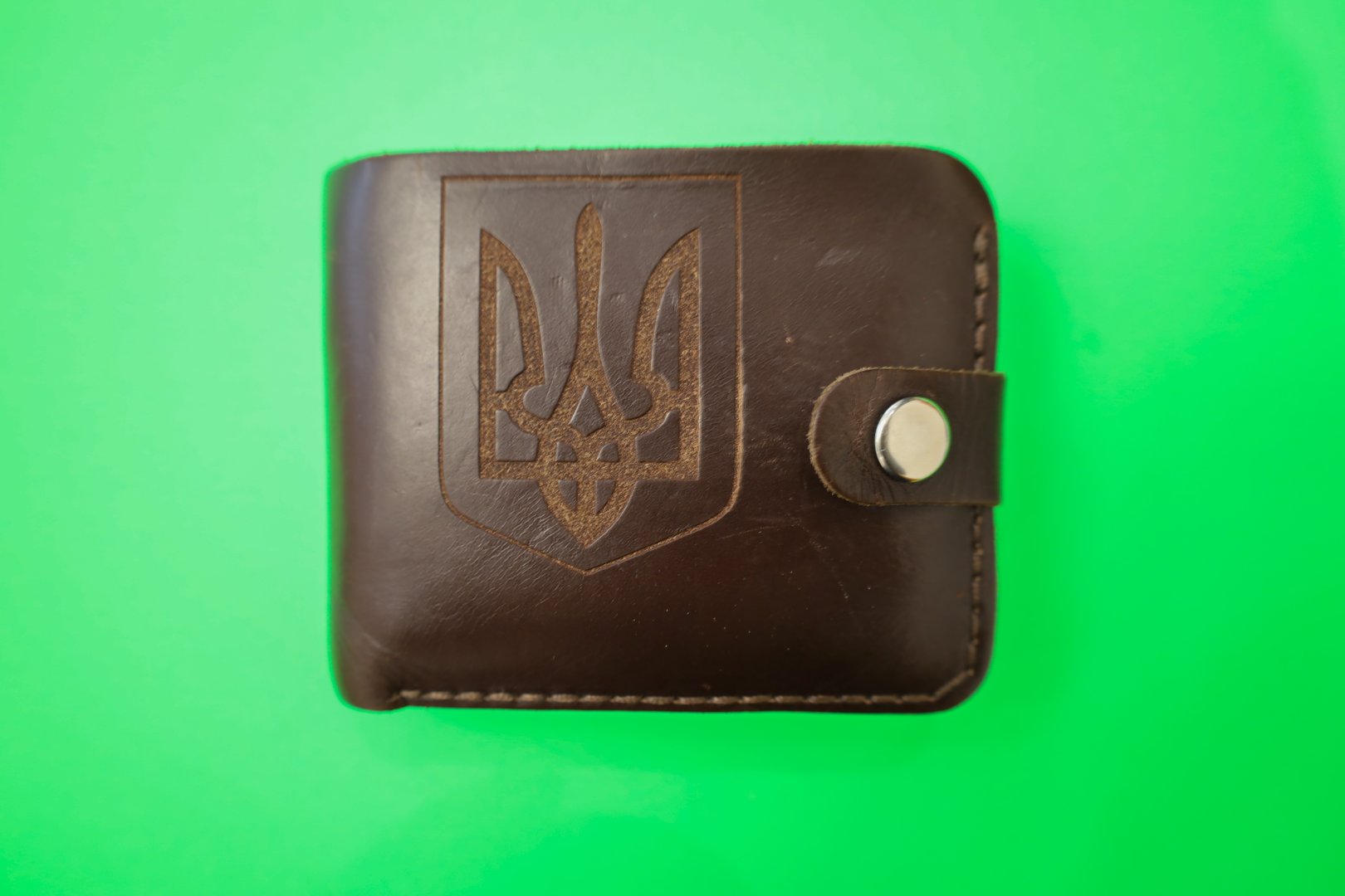 ​A rectangular dark brown leather wallet is adorned with a large engraved Trident (Tryzub), the national coat of arms of Ukraine. Its design is complemented by a neat silver snap-button closure,