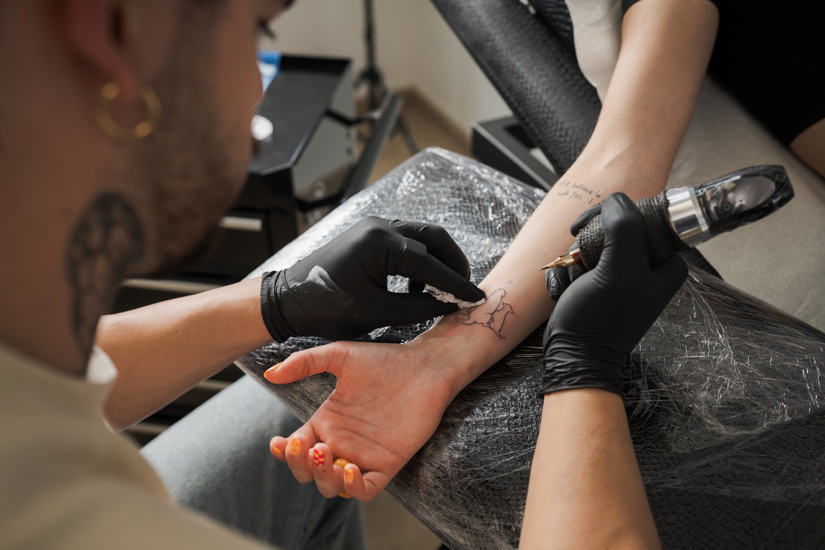 Tattoo artist wearing black gloves is carefully drawing a tattoo on the arm of a client in a professional tattoo studio, cleaning the excess ink with a cotton swab