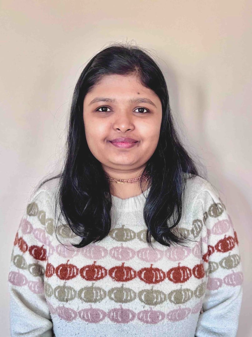 A person with long dark hair wearing a patterned sweater, standing against a plain backdrop.