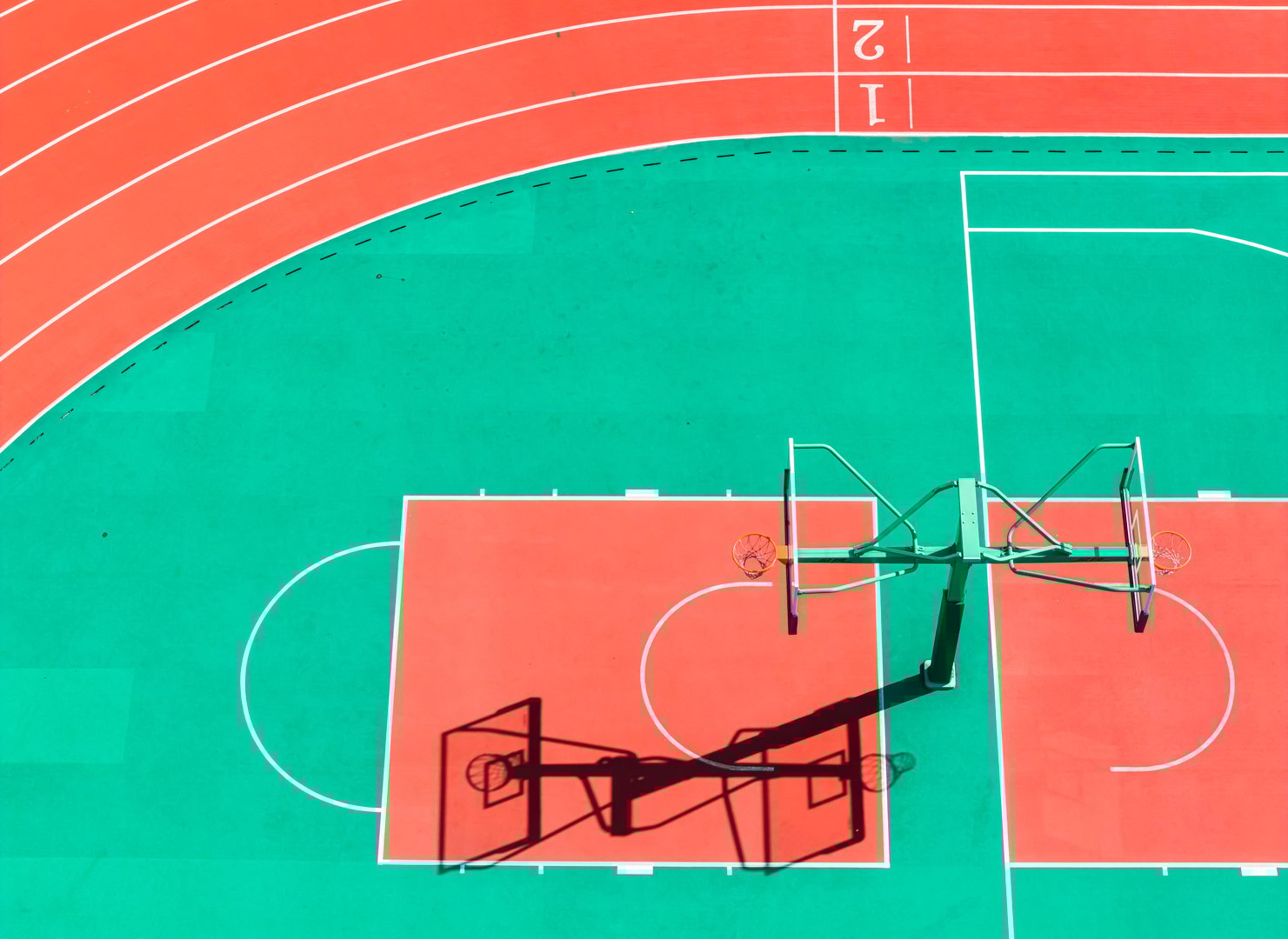 Aerial view of a vibrant basketball court and running track in bold red and green colors, casting distinct shadows.
