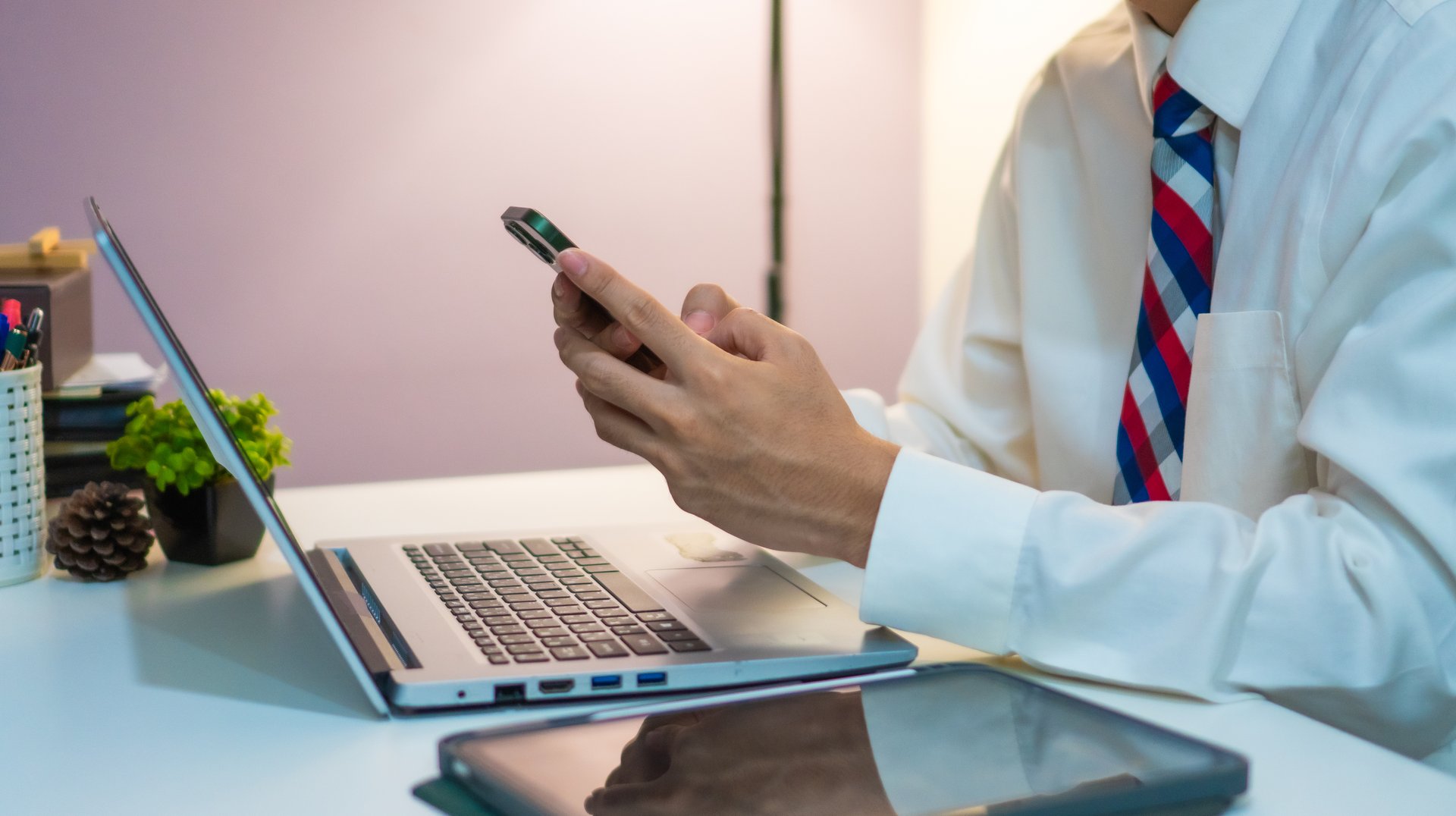 A man in formal attire sits at desk, using smartphone while laptop and tablet are placed nearby, suggesting multitasking and productivity in professional setting
