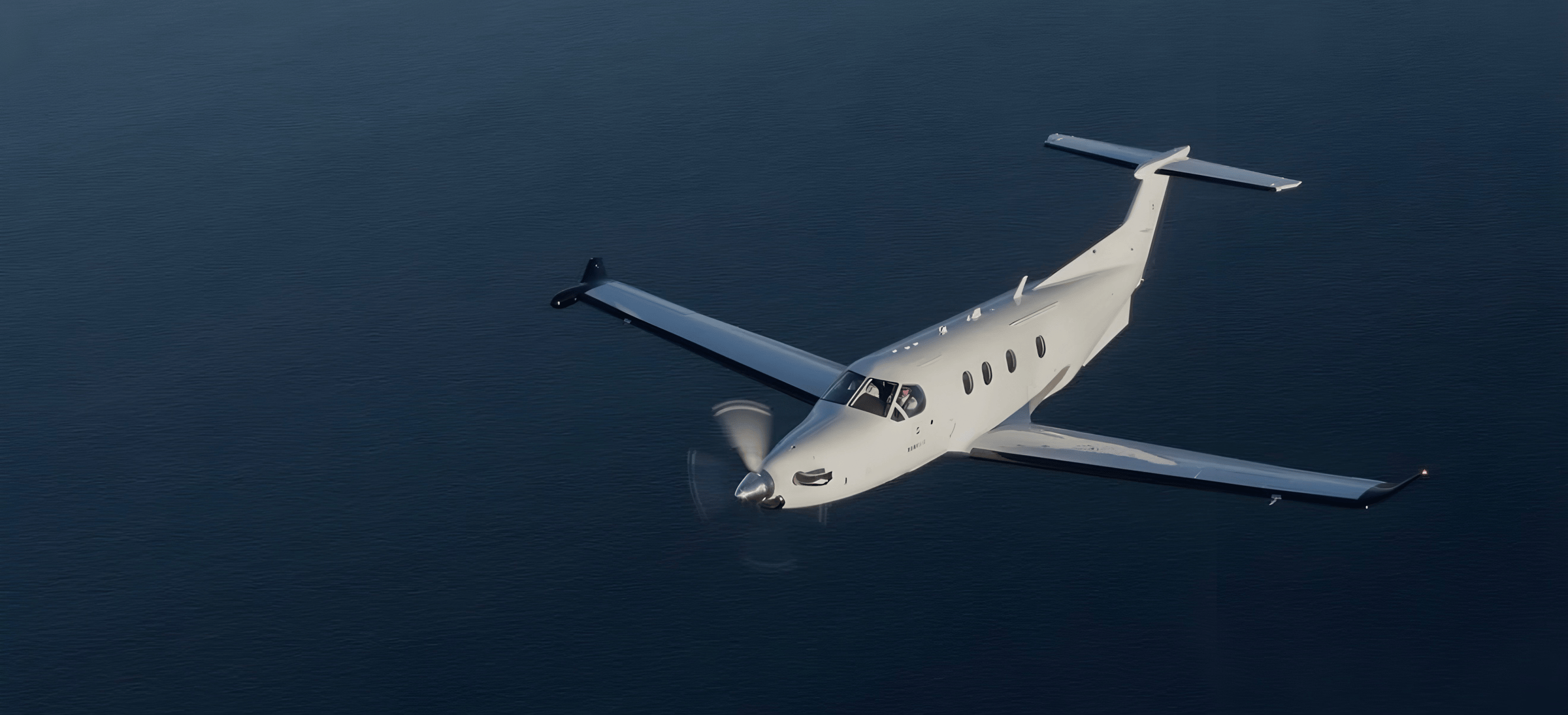 A single-engine propeller plane flying over a large body of dark water, with its landing gear retracted, captured from above.