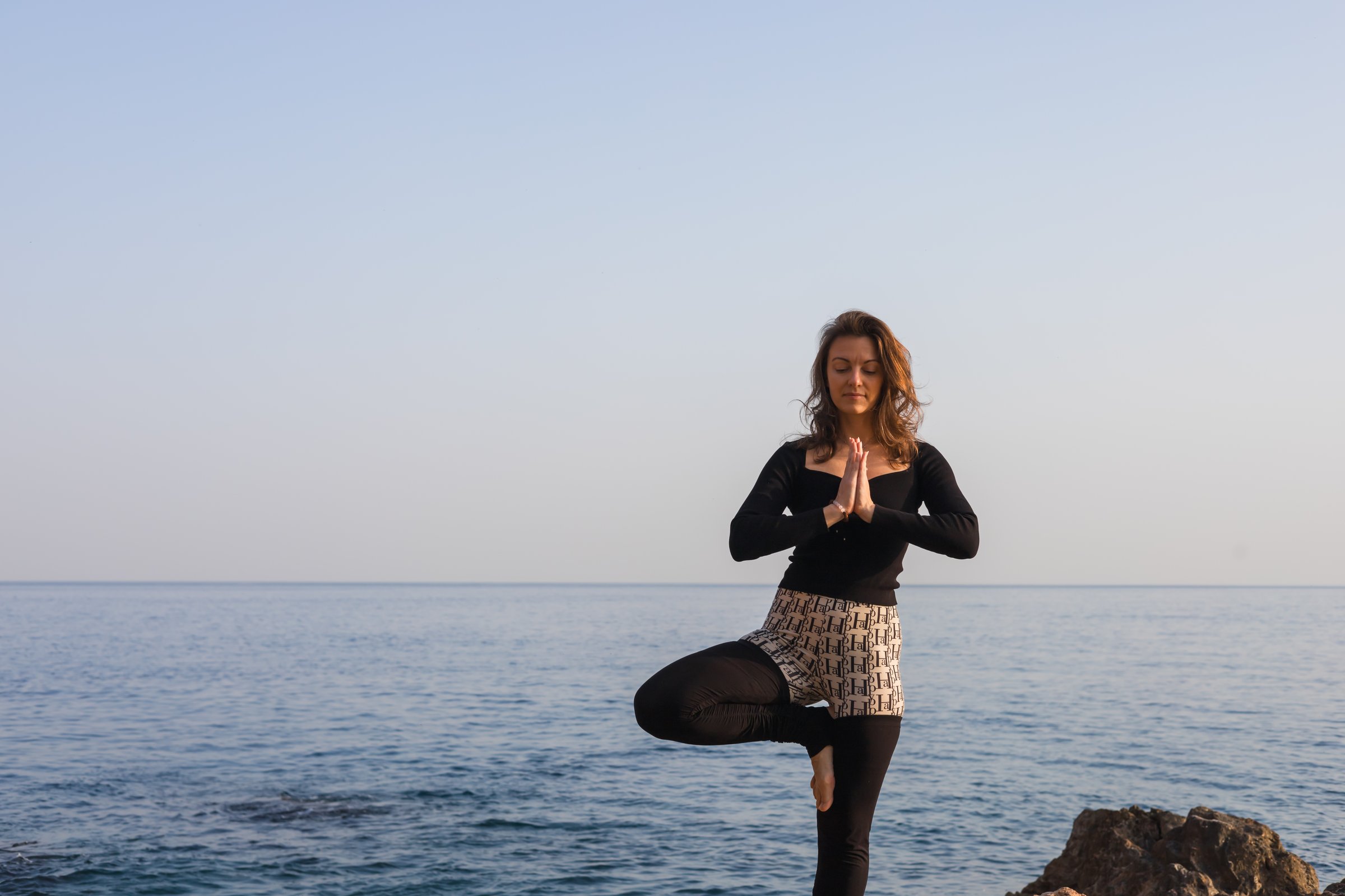 A woman stands on one leg in a yoga pose atop a rocky shoreline. The sun sets over the calm ocean, creating a tranquil atmosphere. Waves gently lap against the rocks below.