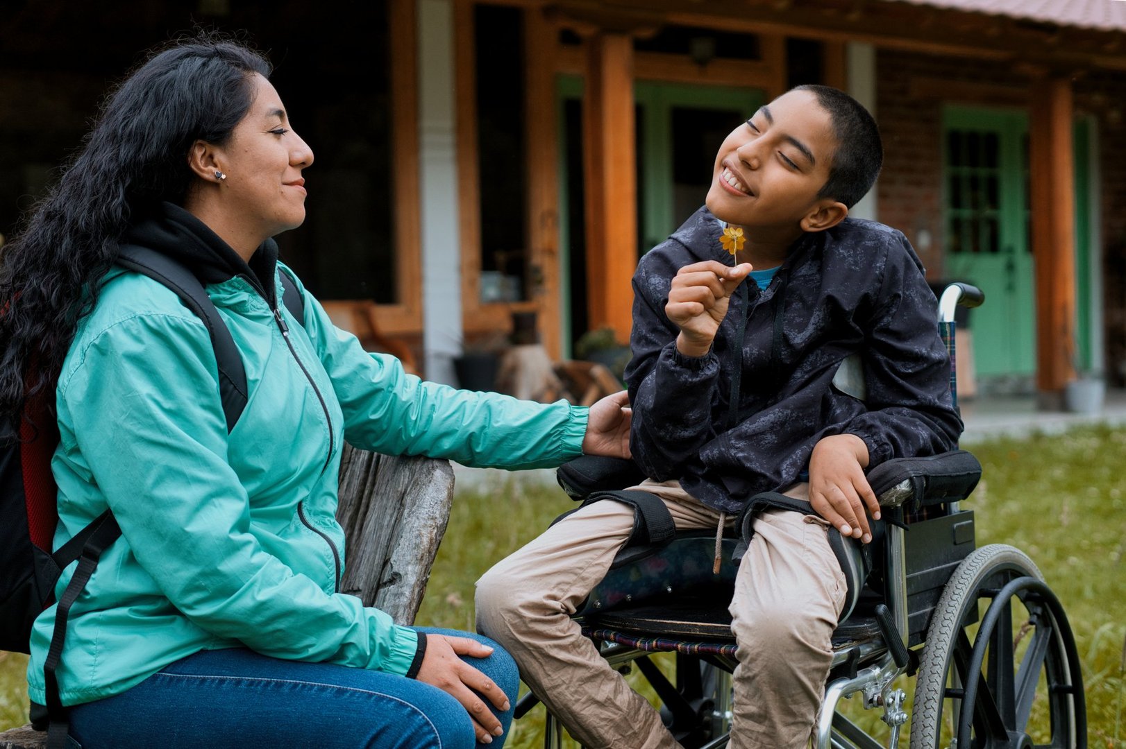 Disabled hispanic child is sitting in his wheelchair, holding a flower and smelling it, while his mother sits next to him and looks at him with love