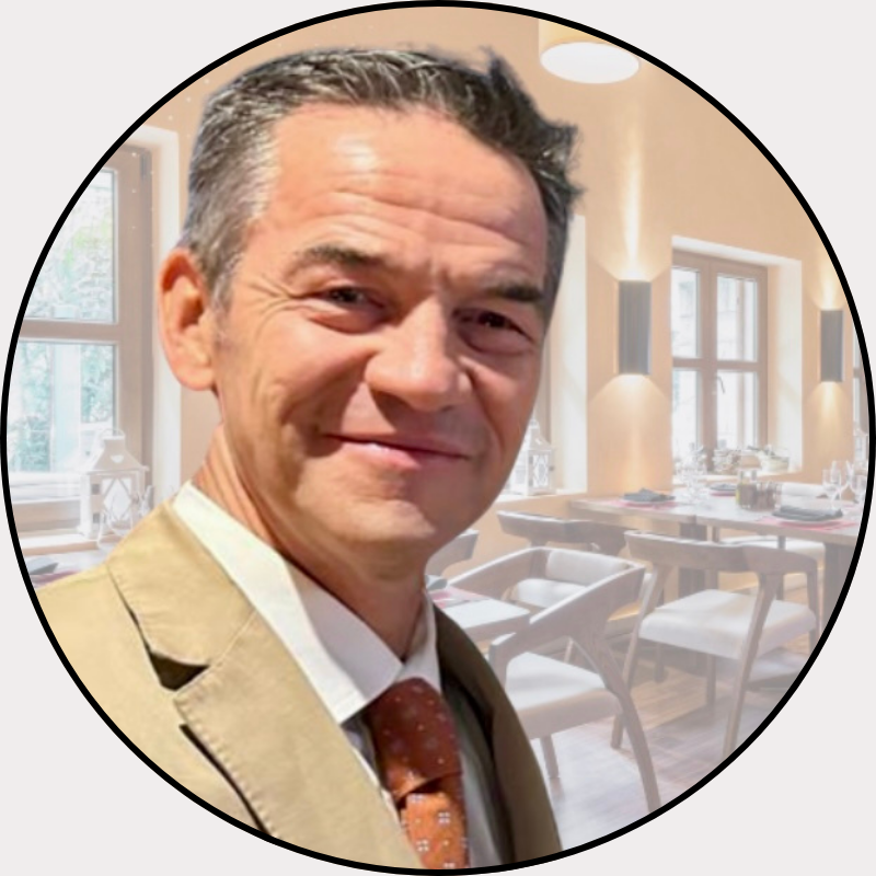 Man in a beige suit and orange tie smiling in a dining room with elegant decor and wooden furniture.