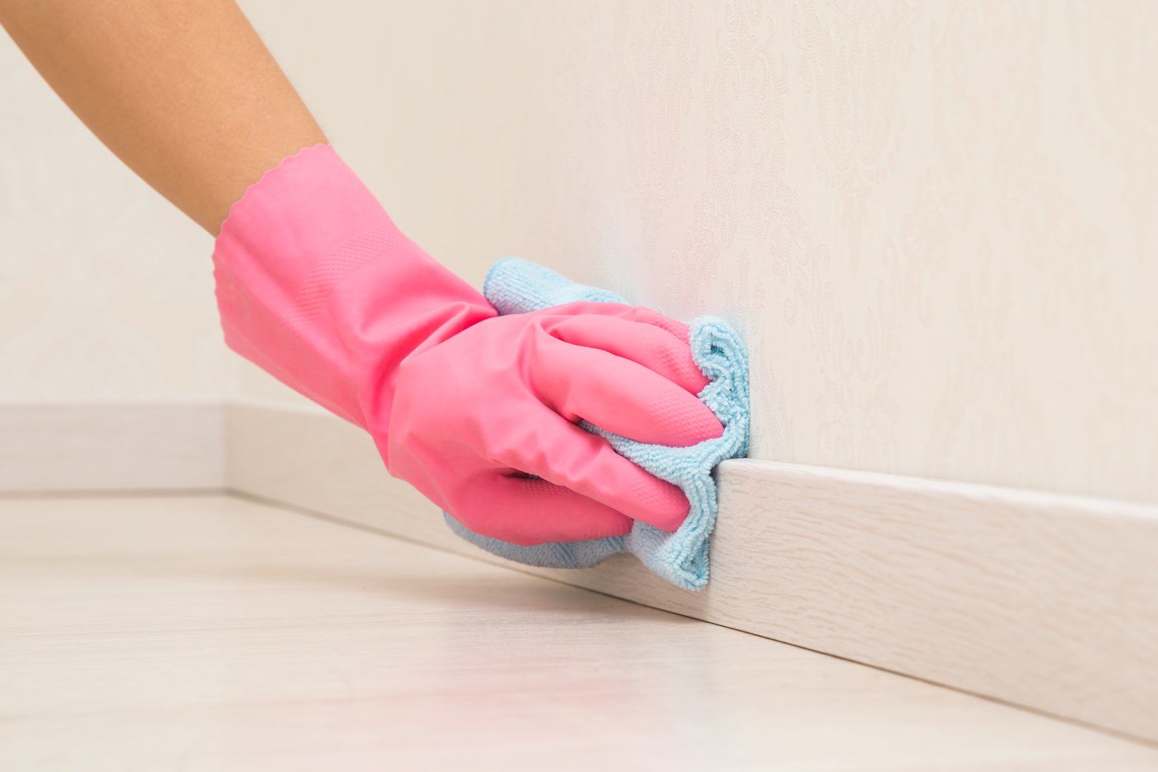 Young adult woman hand in pink rubber protective glove using blue dry rag and wiping light wooden baseboard surface in room at home. Closeup.