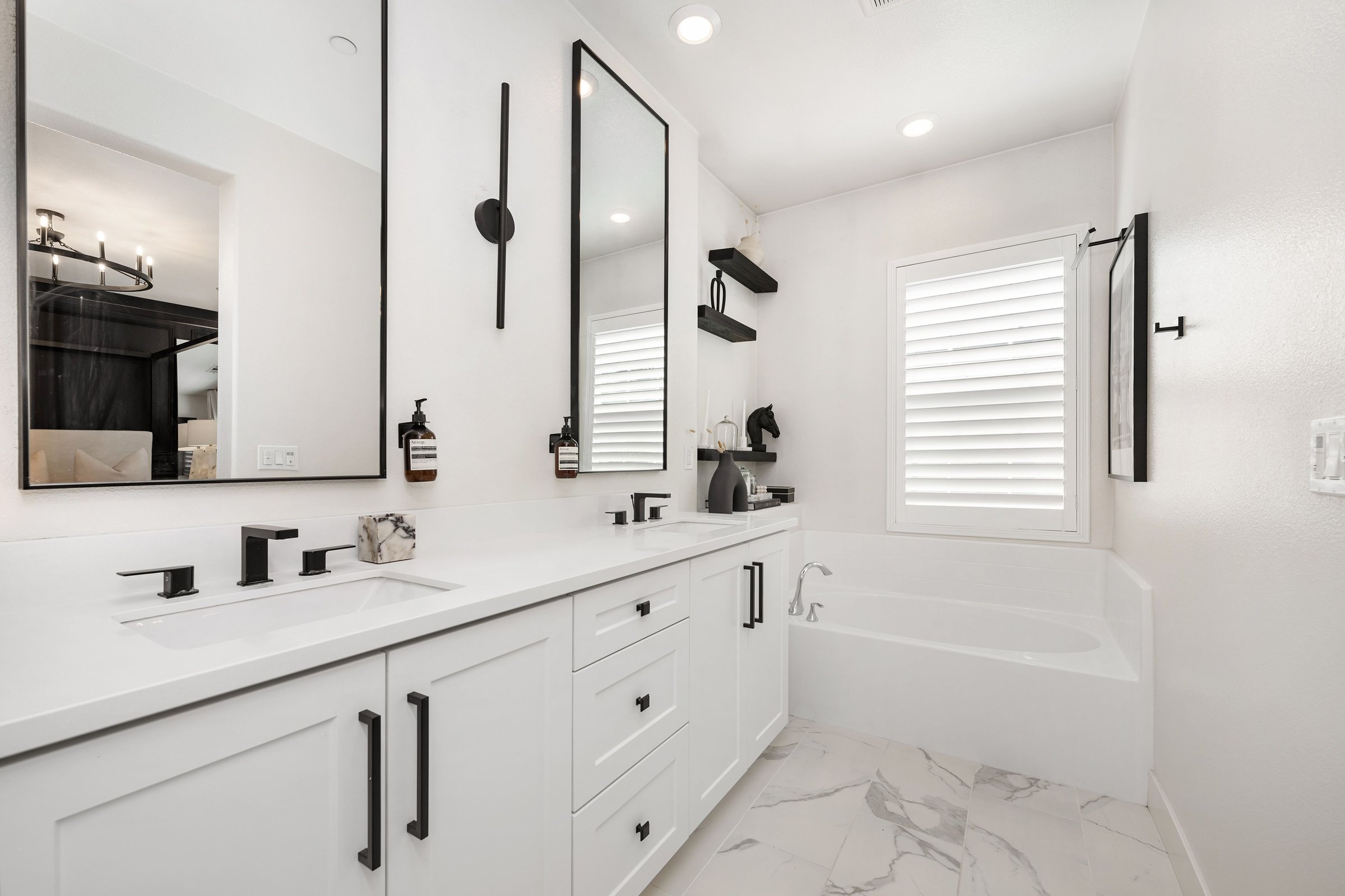 Modern bathroom with white cabinets, black fixtures, and a bathtub under a window with shutters.