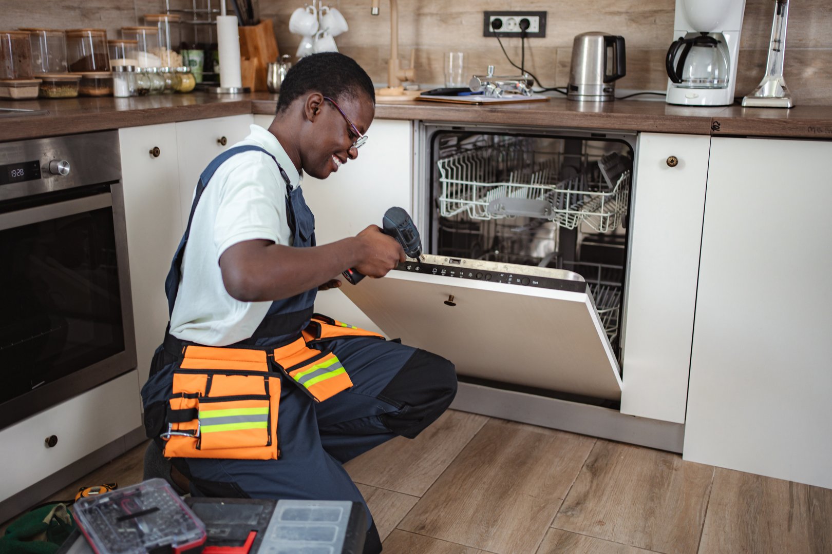 Smiling technician repairing dishwasher using electric screwdriver in modern kitchen