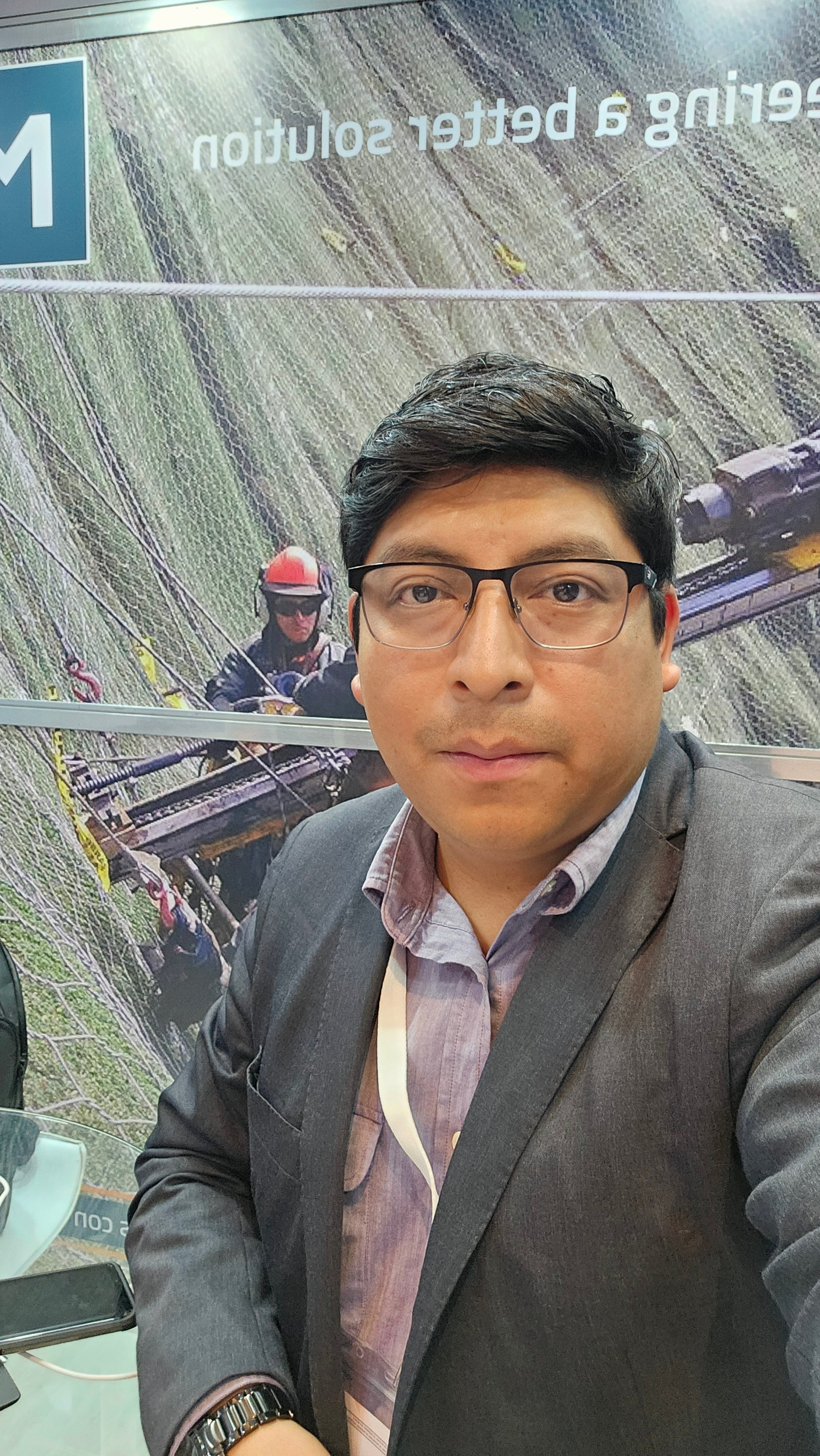 Man in glasses and a suit taking a selfie at an event booth with an industrial background featuring workers and cables.