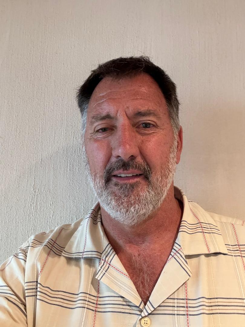 Man with short, dark hair and a beard, wearing a beige and plaid shirt, standing against a plain light background.
