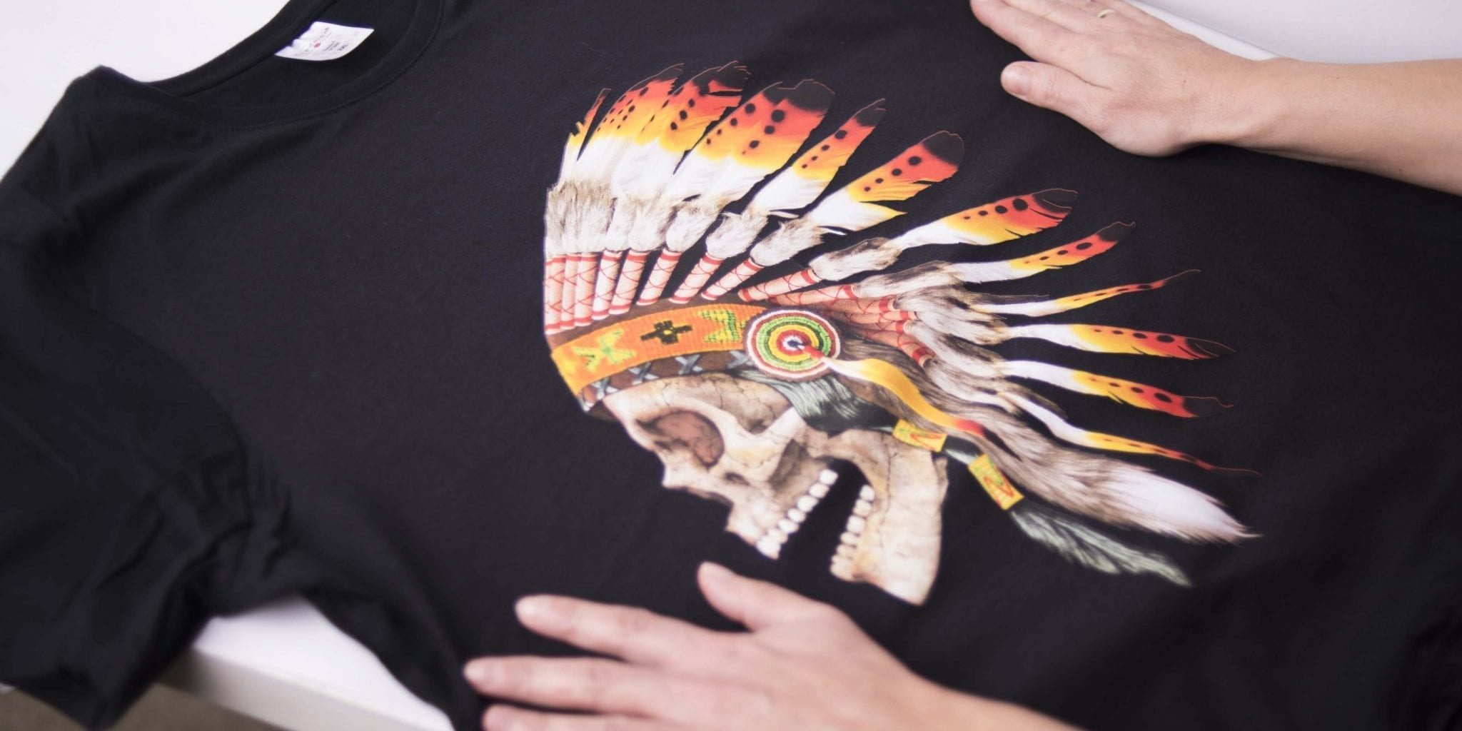 Black t-shirt with a colorful skull wearing a Native American headdress design, held by two hands on a flat surface.