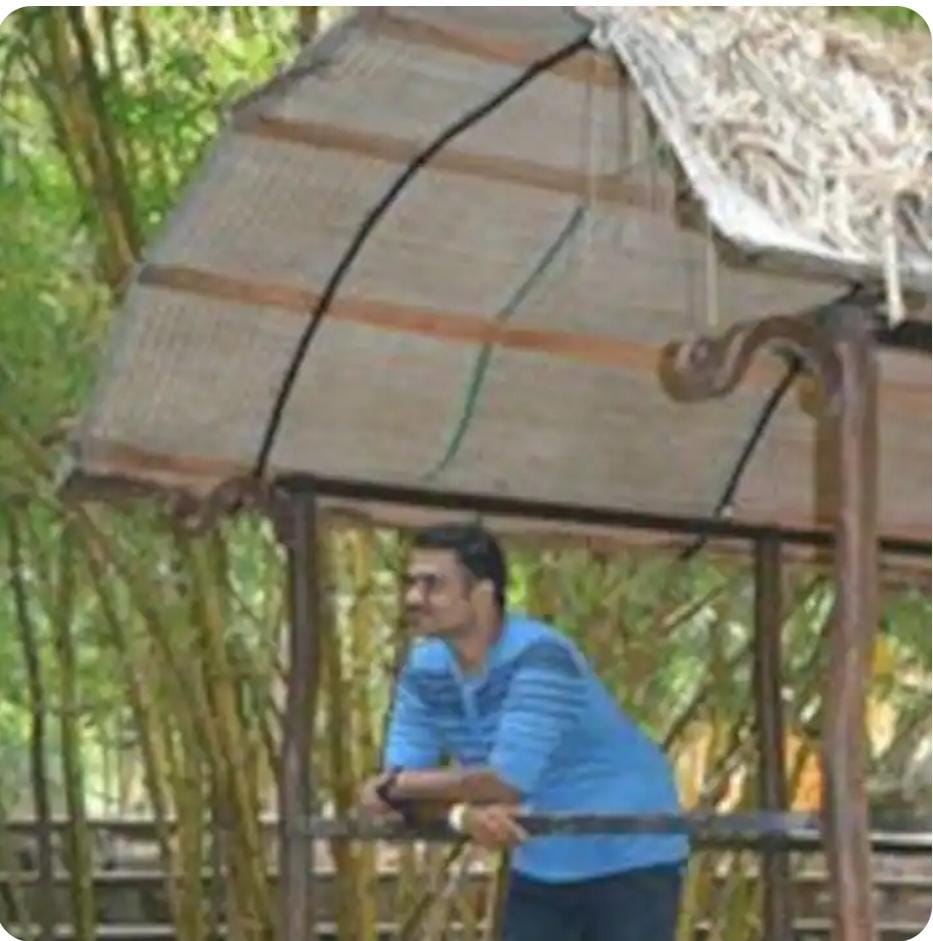 Person in a blue shirt leaning on a wooden railing under a thatched roof with bamboo plants in the background.