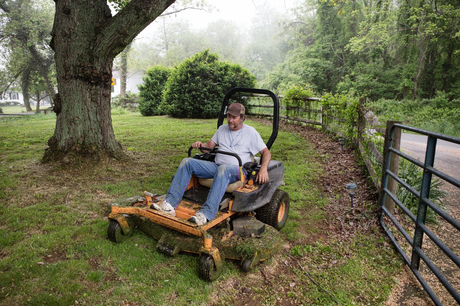 Man cutting grass on a riding lawnmower beside a gate