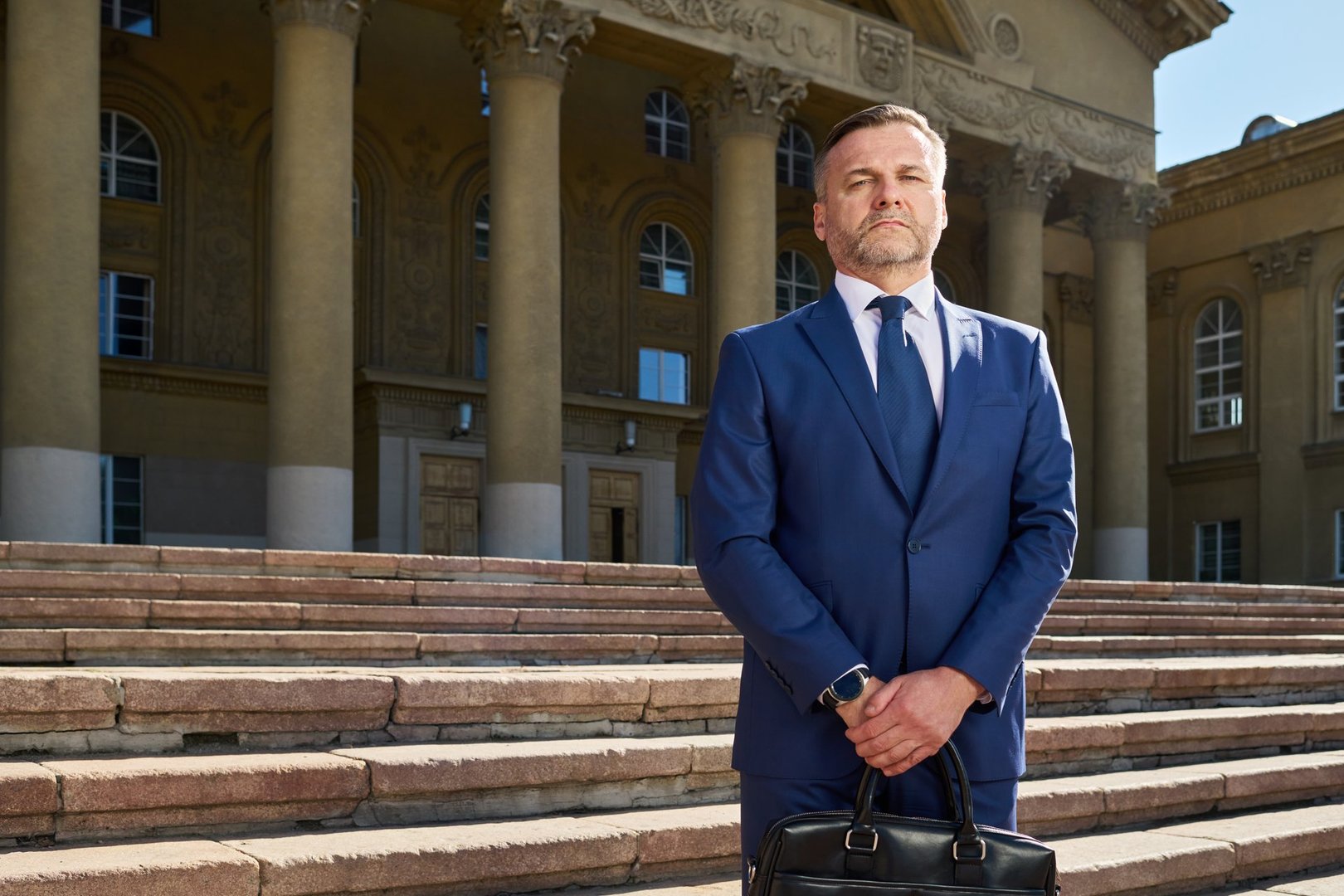 Caucasian middle aged man standing on stone steps in front of classical building holding briefcase wearing business suit looking confidently ahead with serious expression