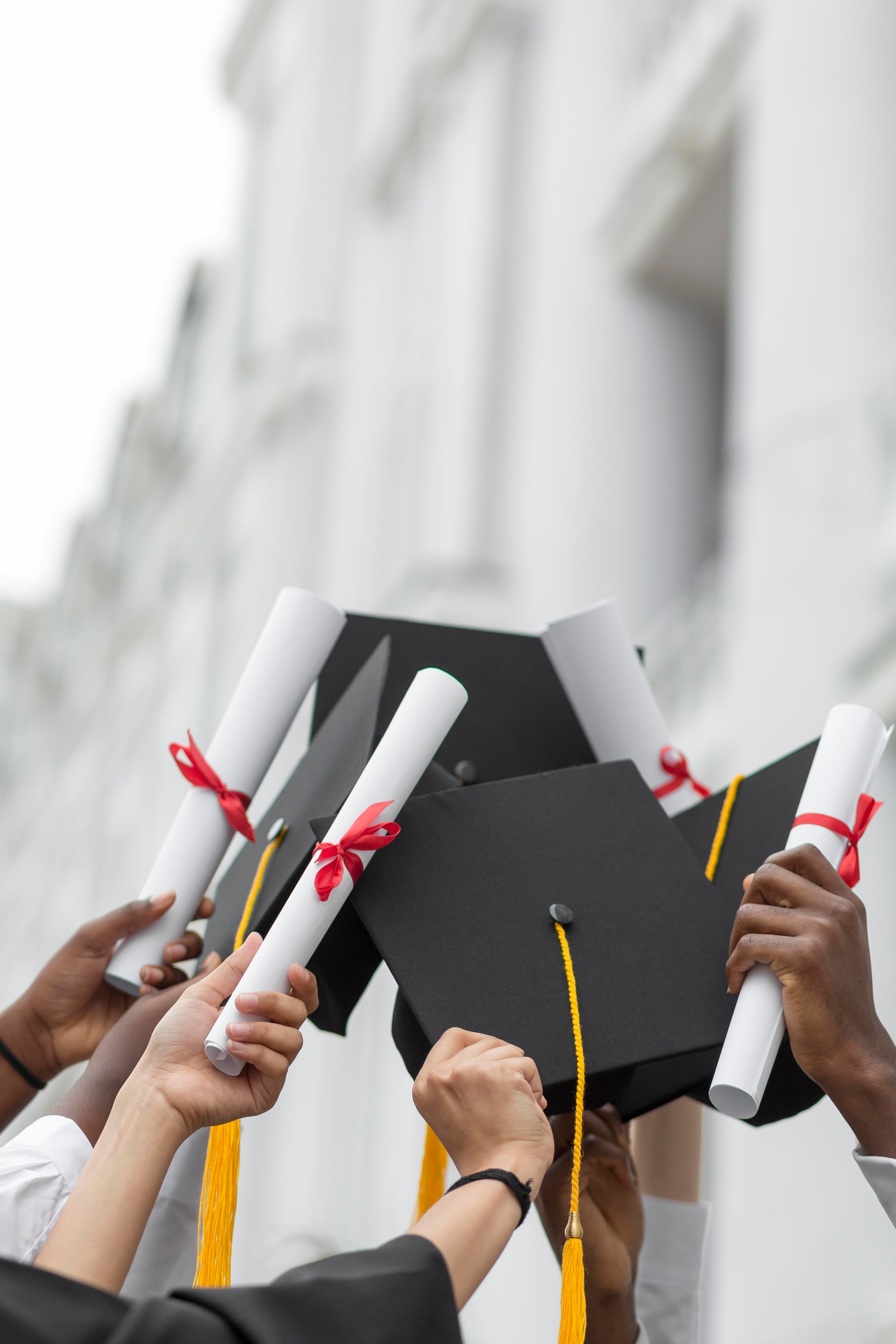 Graduates holding up diplomas and caps, celebrating their achievement, with a blurred building in the background.