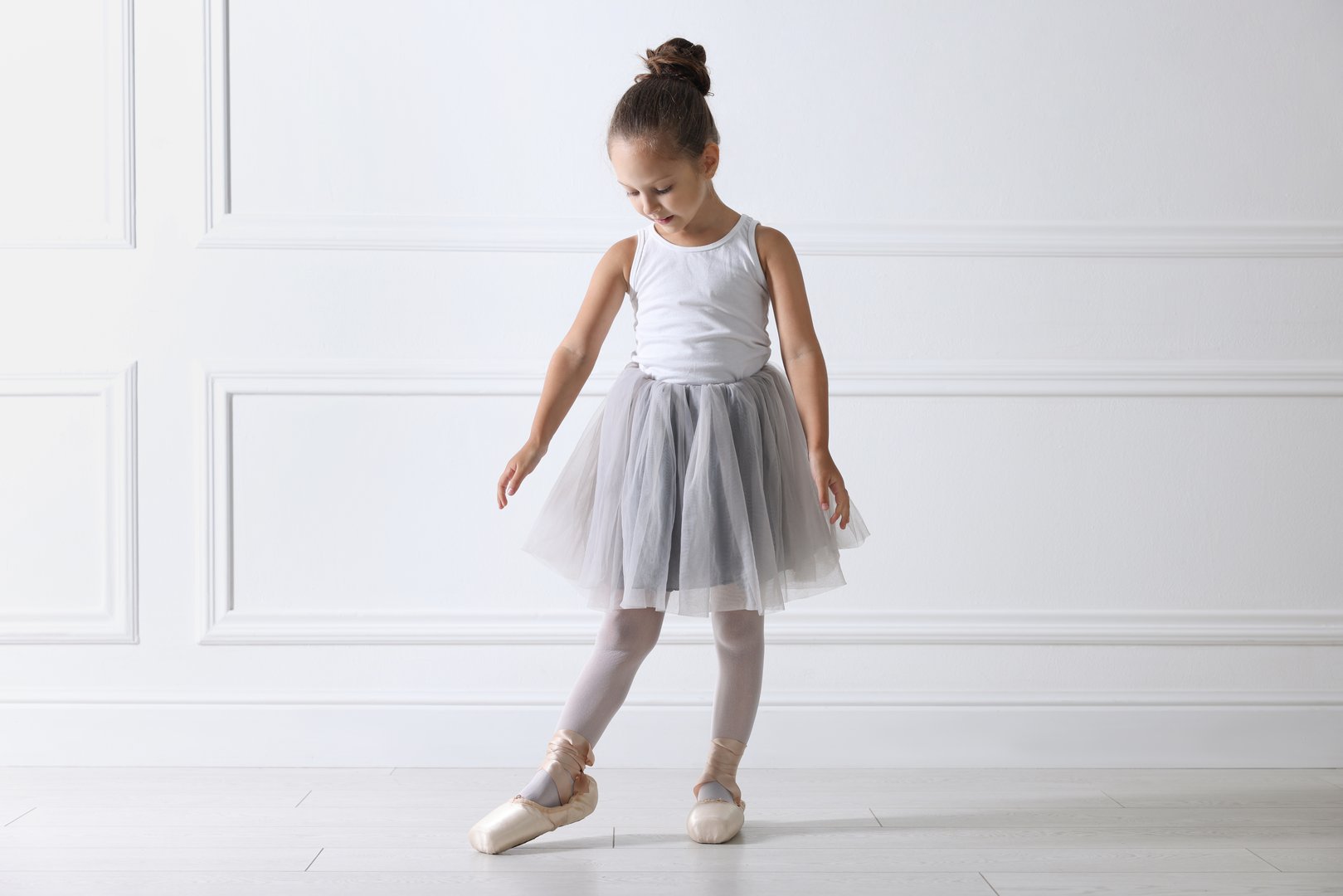 Little ballerina practicing dance moves near white wall indoors