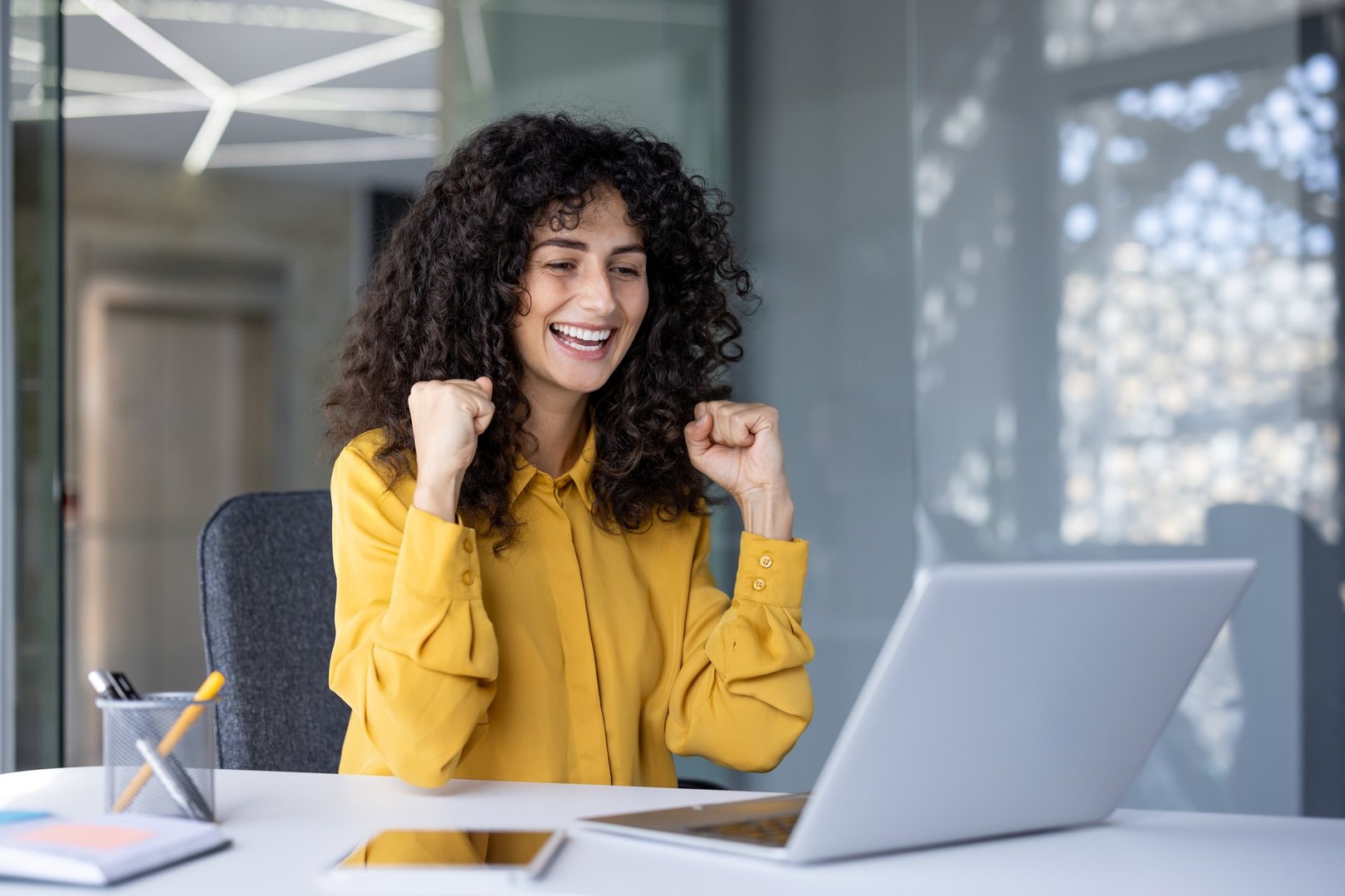 Hispanic businesswoman expresses excitement and success sitting at office desk with laptop. Joyful professional celebrates achievement using technology. Energetic atmosphere positive emotion