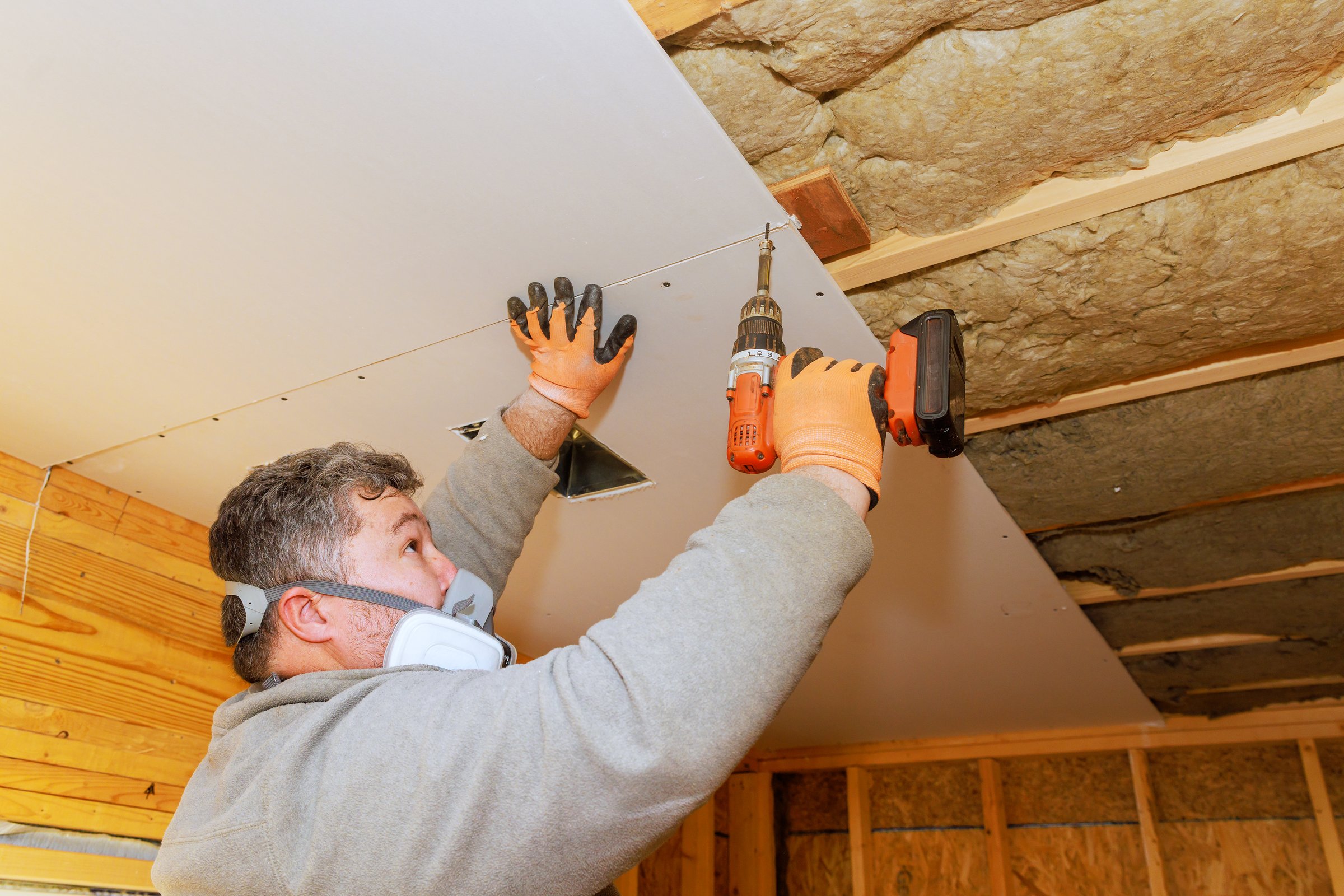 Construction worker secures drywall to ceiling using power drill in cozy indoor area.