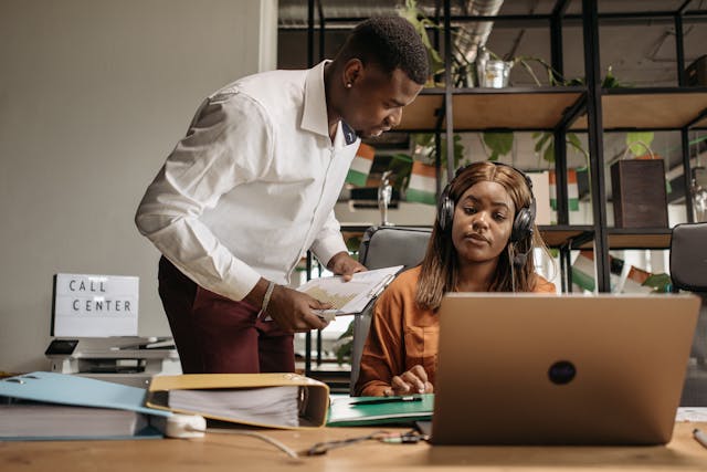 Two people in an office, one wearing headphones at a laptop, the other showing papers. Call center sign in the background.