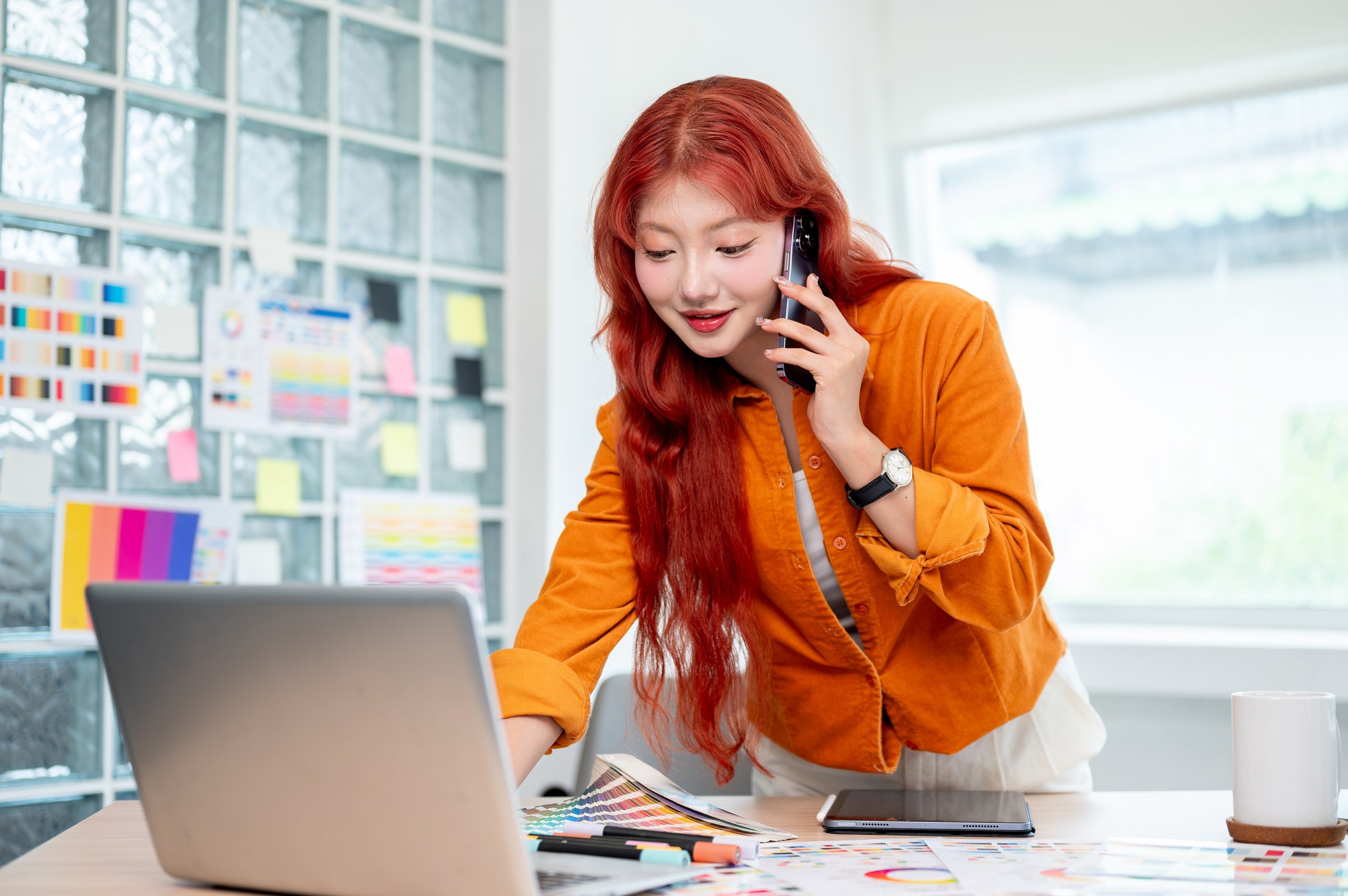 A confident Asian female graphic designer with red curly hair is talking on the phone while leaning against the table and working on her laptop, working in her studio.