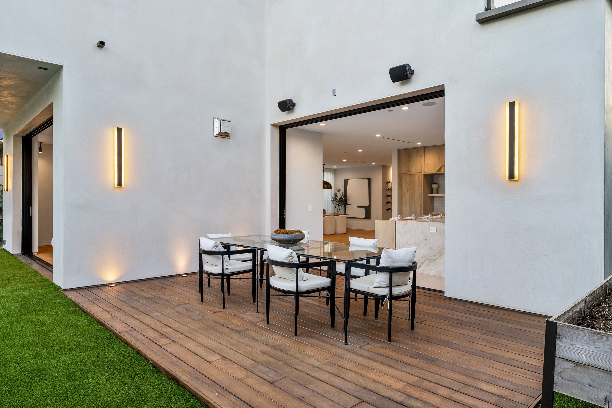 Modern outdoor patio with wooden deck and stylish furniture, adjacent to a well-lit contemporary interior space.