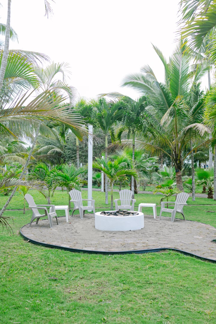 Outdoor seating area with plastic chairs around a fire pit, surrounded by lush palm trees.