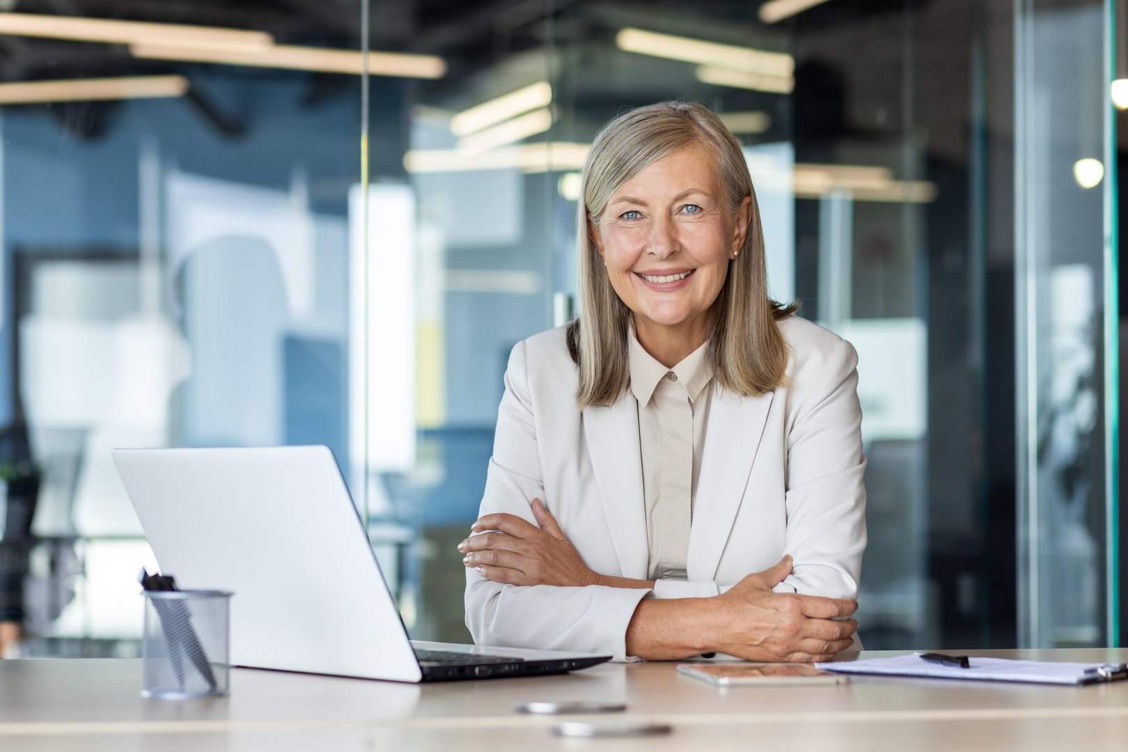 Portrait of senior mature gray-haired businesswoman inside office, female boss sitting at desk and looking at camera with crossed arms, experienced financier with laptop at workplace.