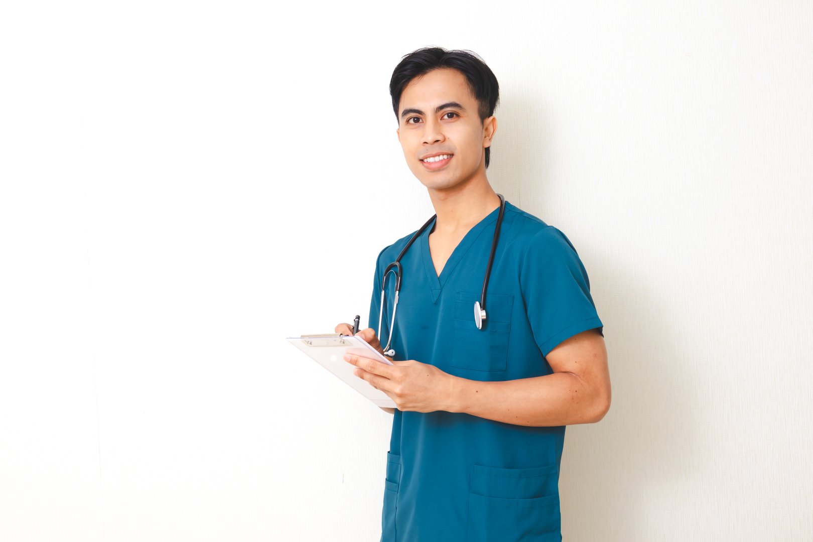 Smiling young male nurse in medical scrubs with stethoscope holding clipboard, confident healthcare professional portrait