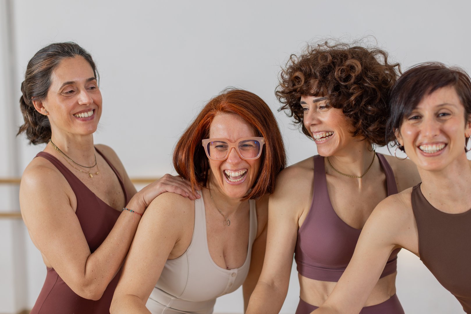 Four diverse women laughing and leaning together in a fitness studio. Ideal for concepts like female friendship, wellness, positive energy and active lifestyle. Horizontal, medium close-up.