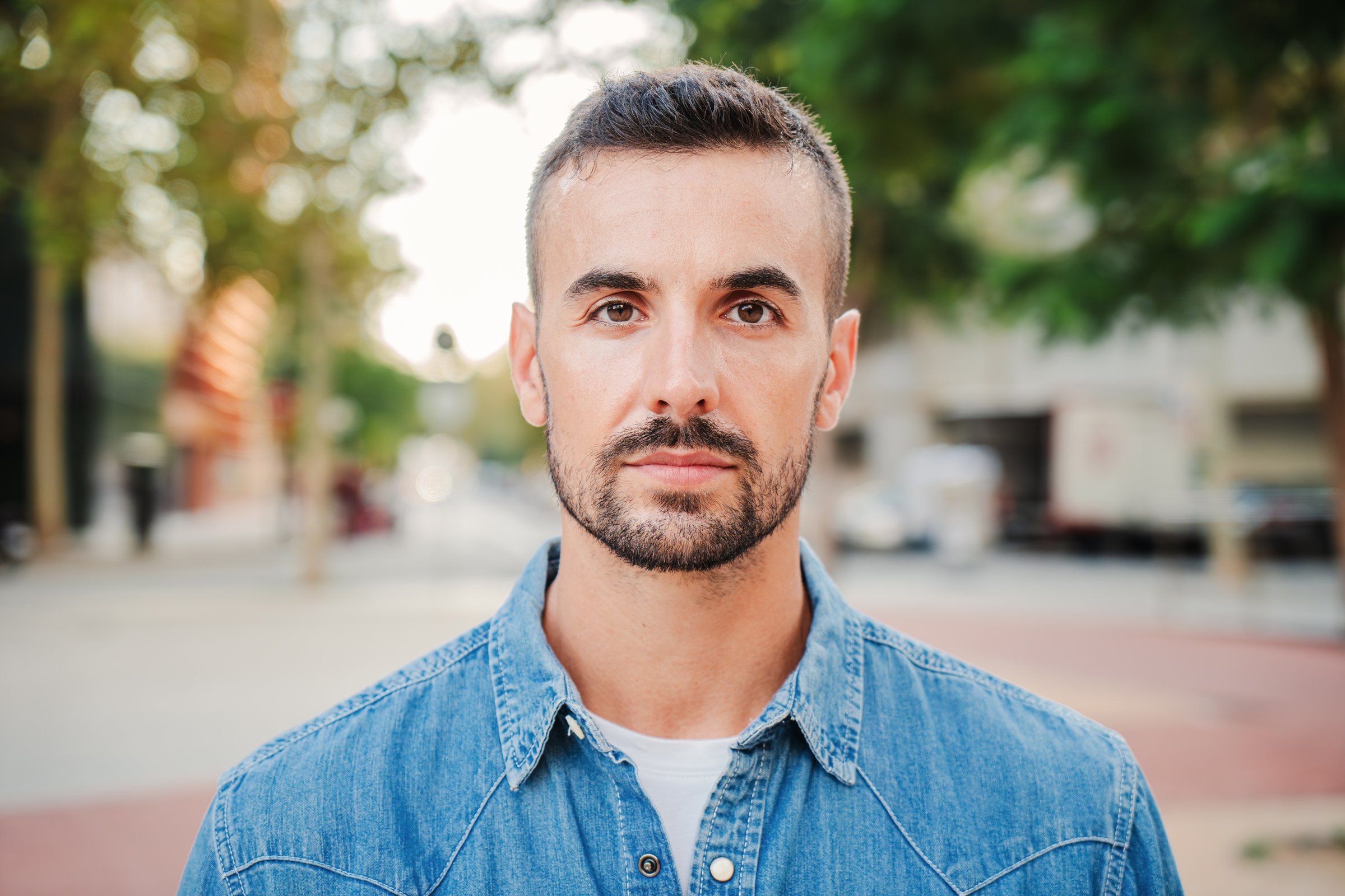 Close up individual portrait of handsome serious guy looking at camera standing outdoors. Front view of young real man with pensive expression. Head shot of one formal adult male staring thoughtful. High quality photo