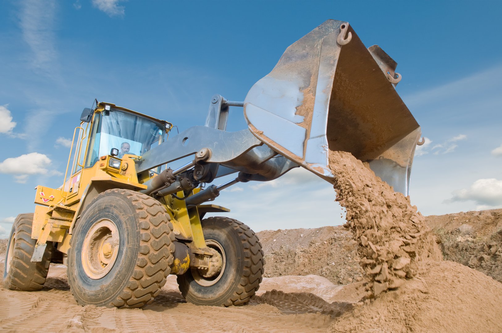 A yellow bulldozer lifting and dumping sand on a construction site under a clear blue sky.