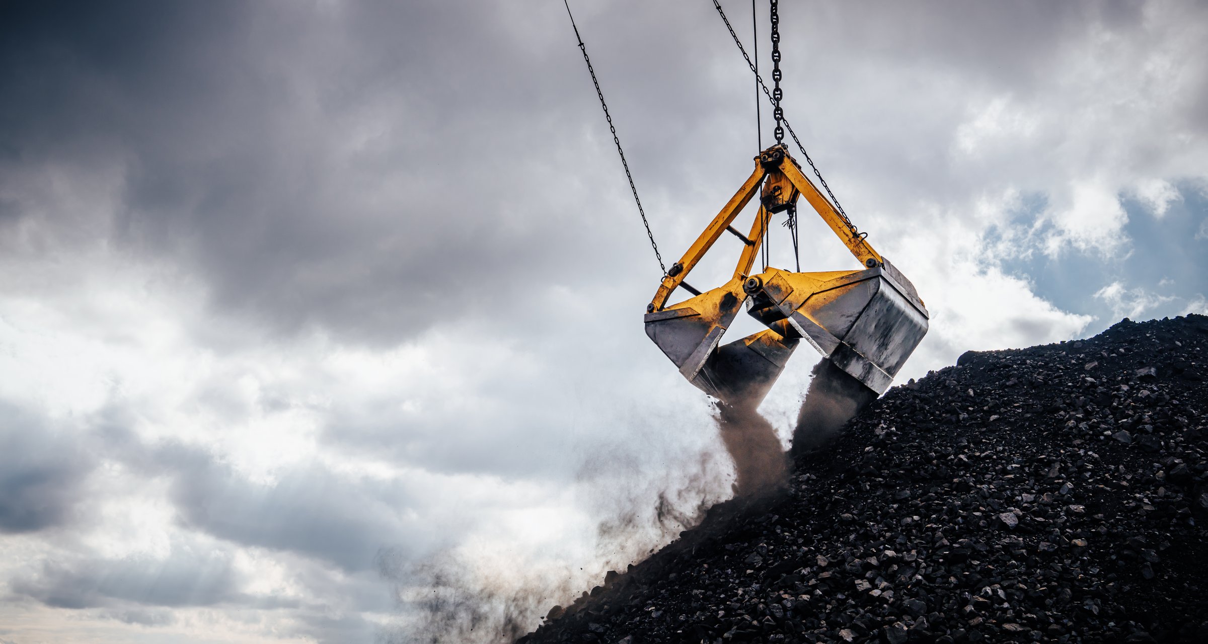 An excavator crane is engaged in loading coal using a large bucket, which is suspended and poised over a mound of black coal. Dust is billowing from the bucket as it lifts, indicating the heavy load being transported. The cloudy sky