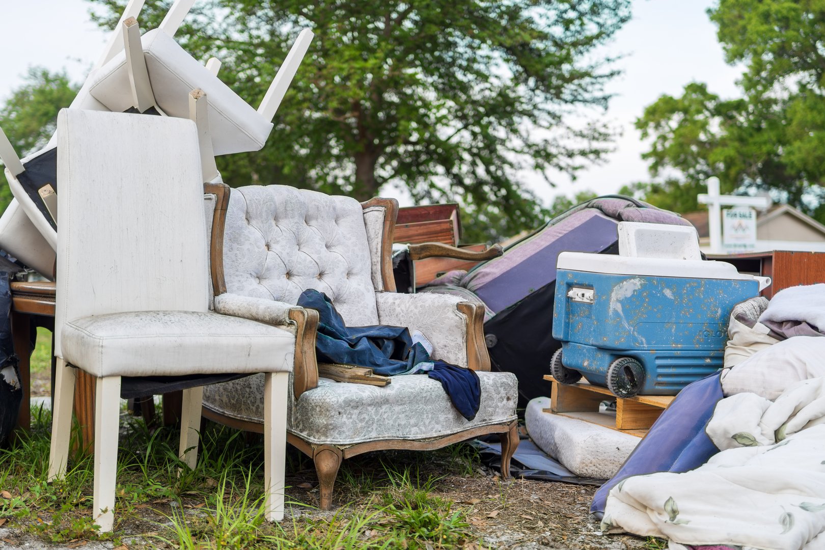 A cluttered collection of household items, including upholstered chairs and a vintage cooler, sits abandoned on a suburban curb