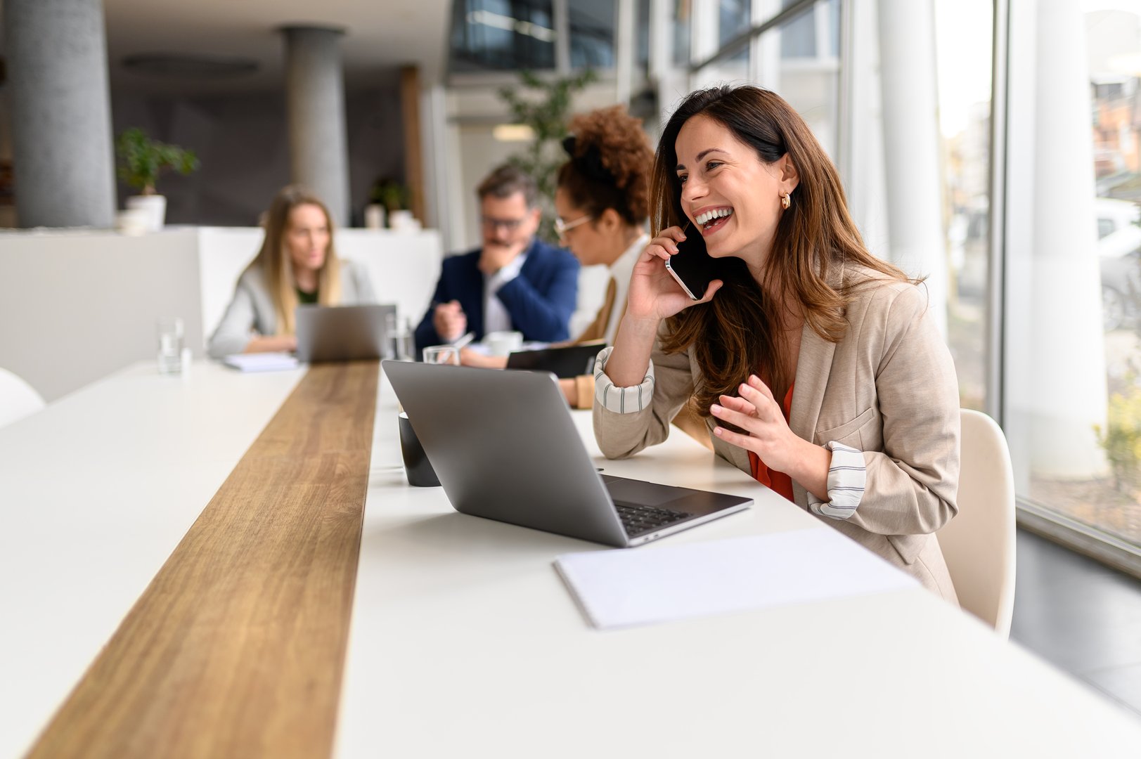 Cheerful businesswoman talking over smart phone while sitting with laptop at conference table during business meeting