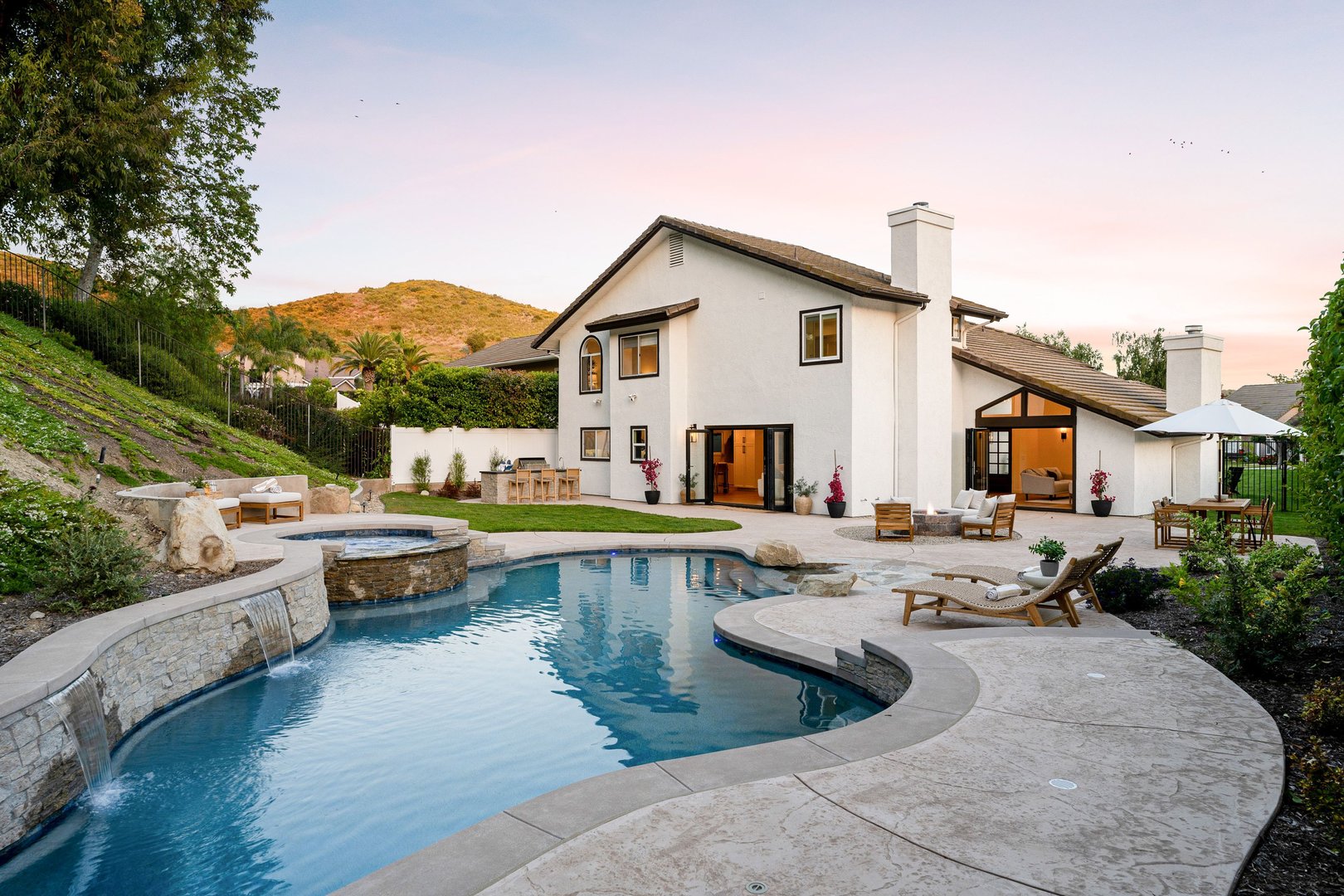 A luxurious backyard with a swimming pool, hot tub, and patio furniture at a modern house during sunset.