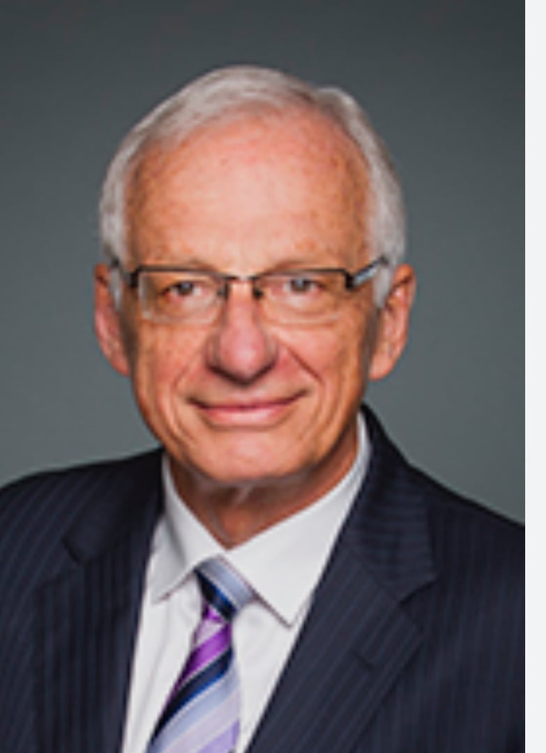 Elderly man with glasses, wearing a suit and striped tie, smiling slightly against a gray background.
