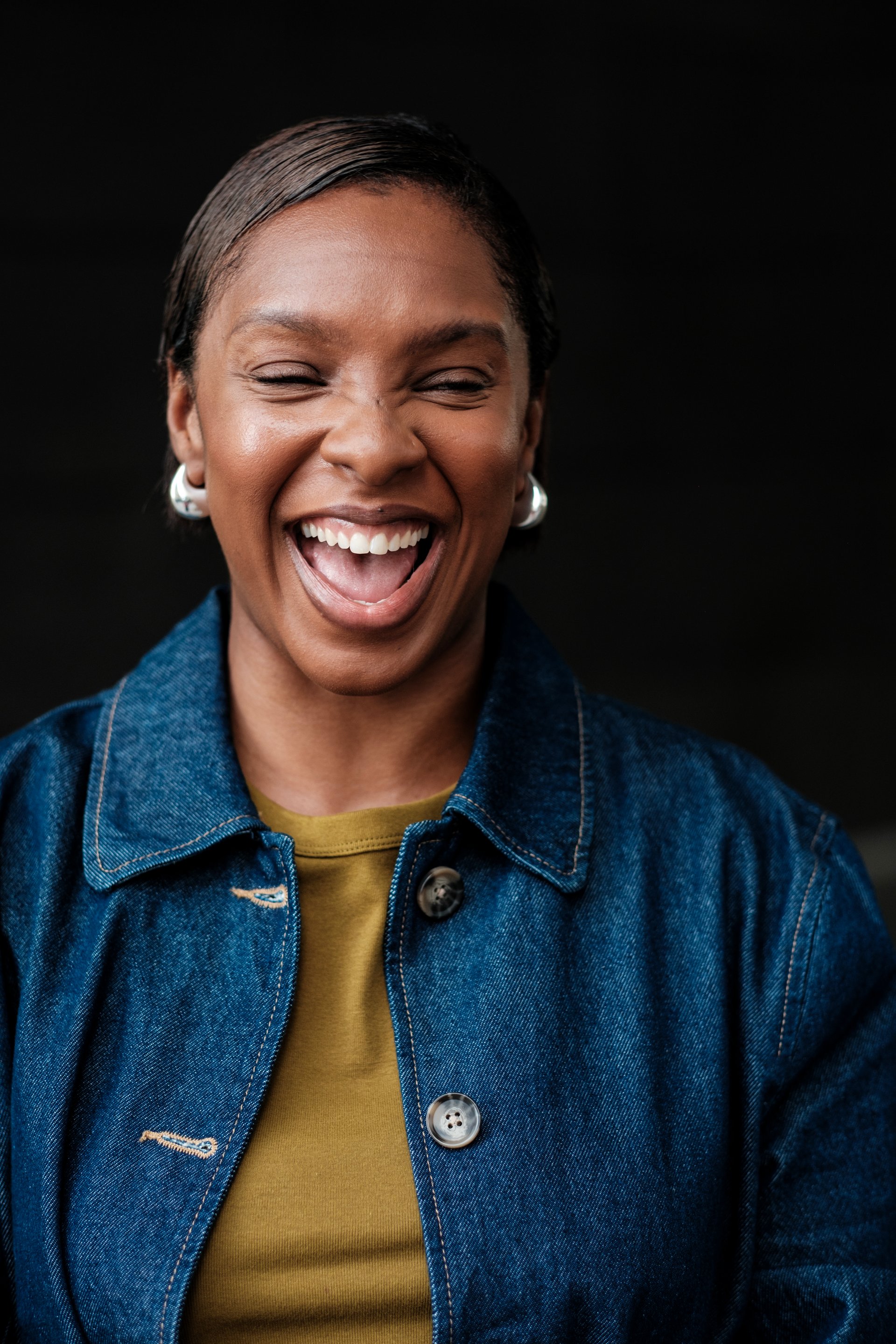 Studio portrait of a cheerful mid adult black woman with short hair, wearing a denim jacket and a khaki t-shirt, laughing with eyes closed on a black background