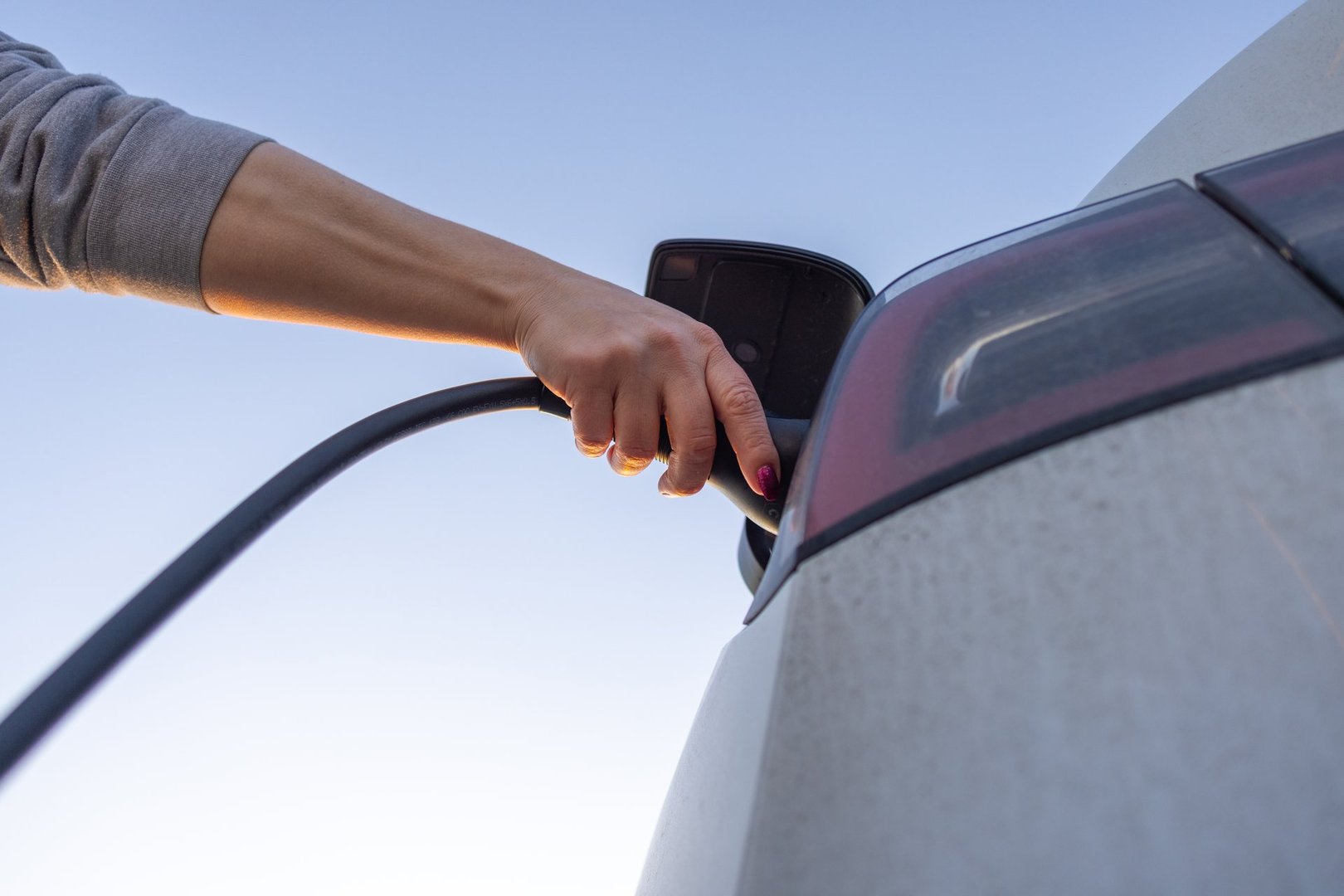 A close-up of a person plugging a charger into an electric car with a clear blue sky in the background. This image symbolizes eco-friendly transportation, sustainable energy solutions, and the shift toward renewable energy in modern mobility.