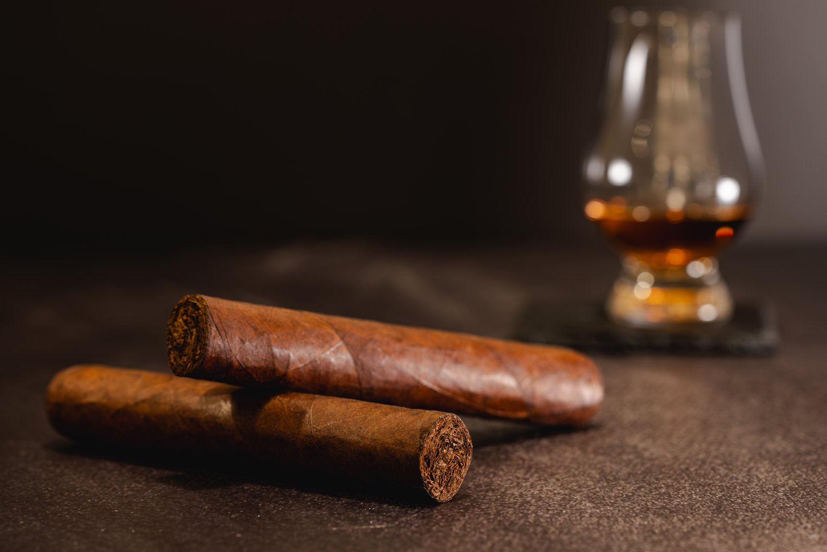 Premium hand-rolled cigars on a dark, textured surface, eloquently accompanied by a glass of aged, golden rum in the soft-focus background, evoking a sense of luxury and relaxation. Perfect for connoisseurs of fine tobacco and spirits.