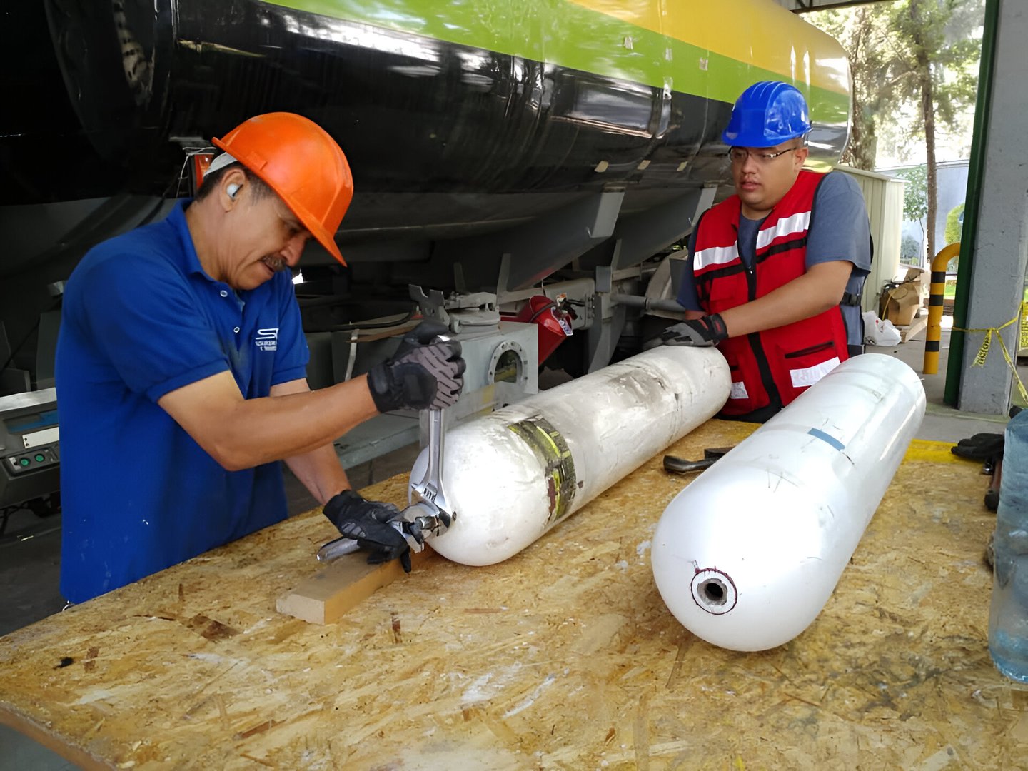Two workers with helmets inspect and prepare large white gas cylinders on a wooden table near industrial machinery.
