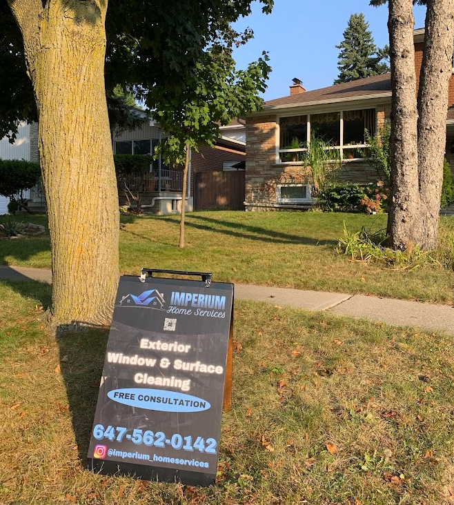 A sign for Imperium Home Services offers exterior cleaning services, placed on a lawn in front of a house on a sunny day.